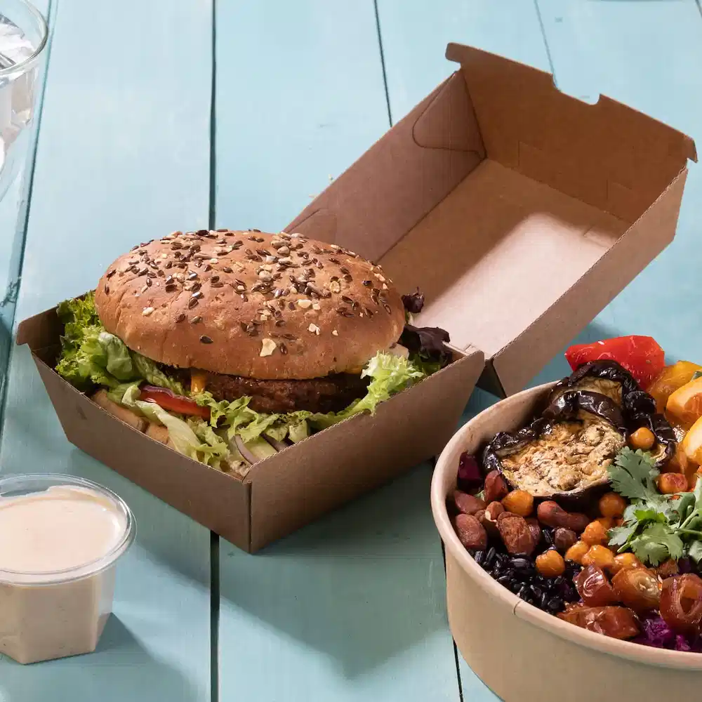 Un burger con panino ai semi di sesamo e lattuga in un box da asporto marrone, accanto a una ciotola di verdure e legumi misti.