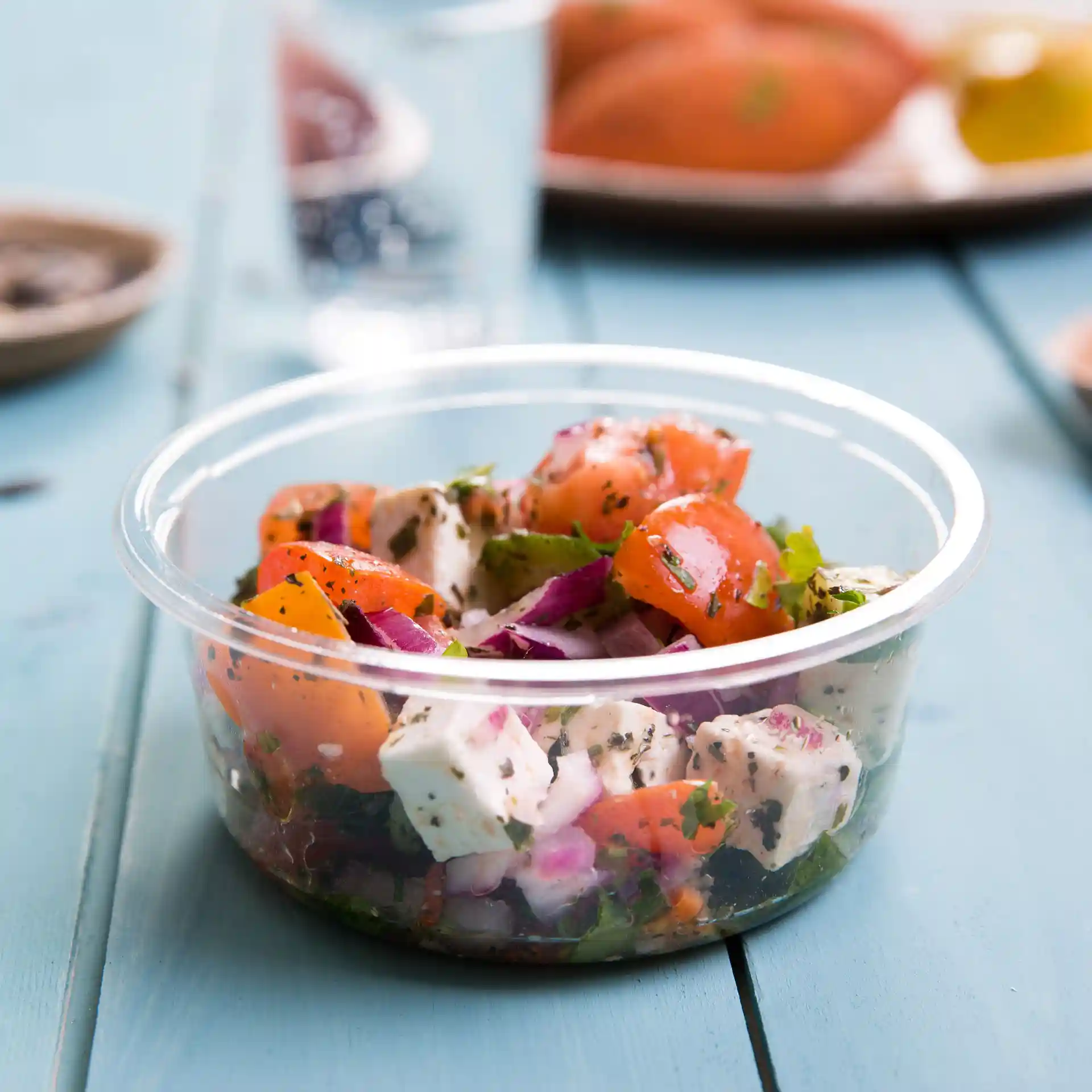 Contenitore rotondo trasparente con un mix di insalata di pomodori, cipolle e erbe, posato su una superficie in legno blu.