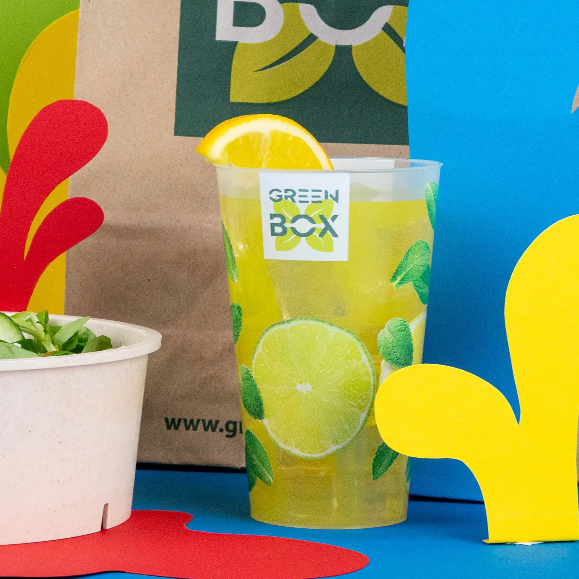 Bicchiere riutilizzabile trasparente con logo, riempito con una bevanda gialla e guarnito con una fetta di limone, accanto a una ciotola di insalata.