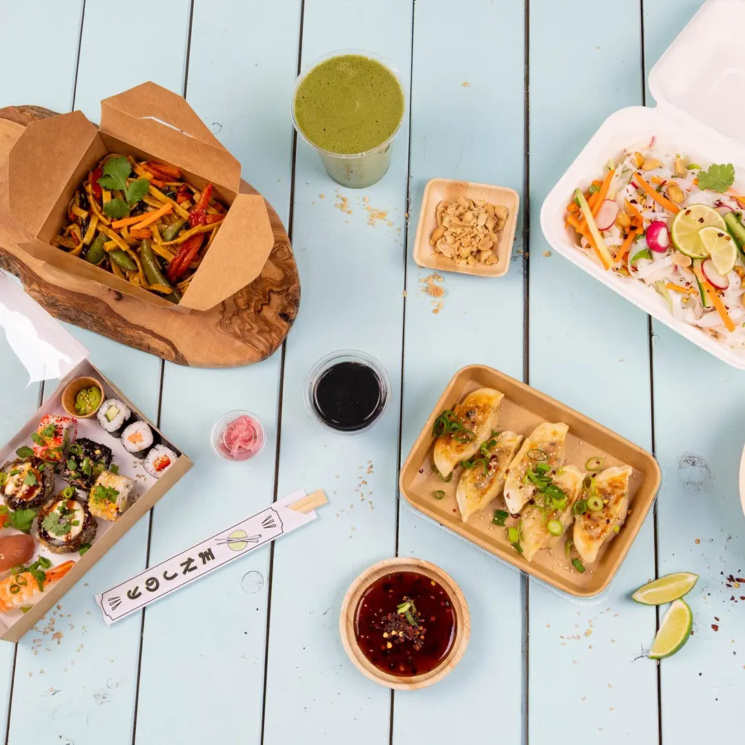 Piatti asiatici assortiti tra cui sushi, ravioli, verdure saltate e una bevanda verde su un tavolo di legno blu.