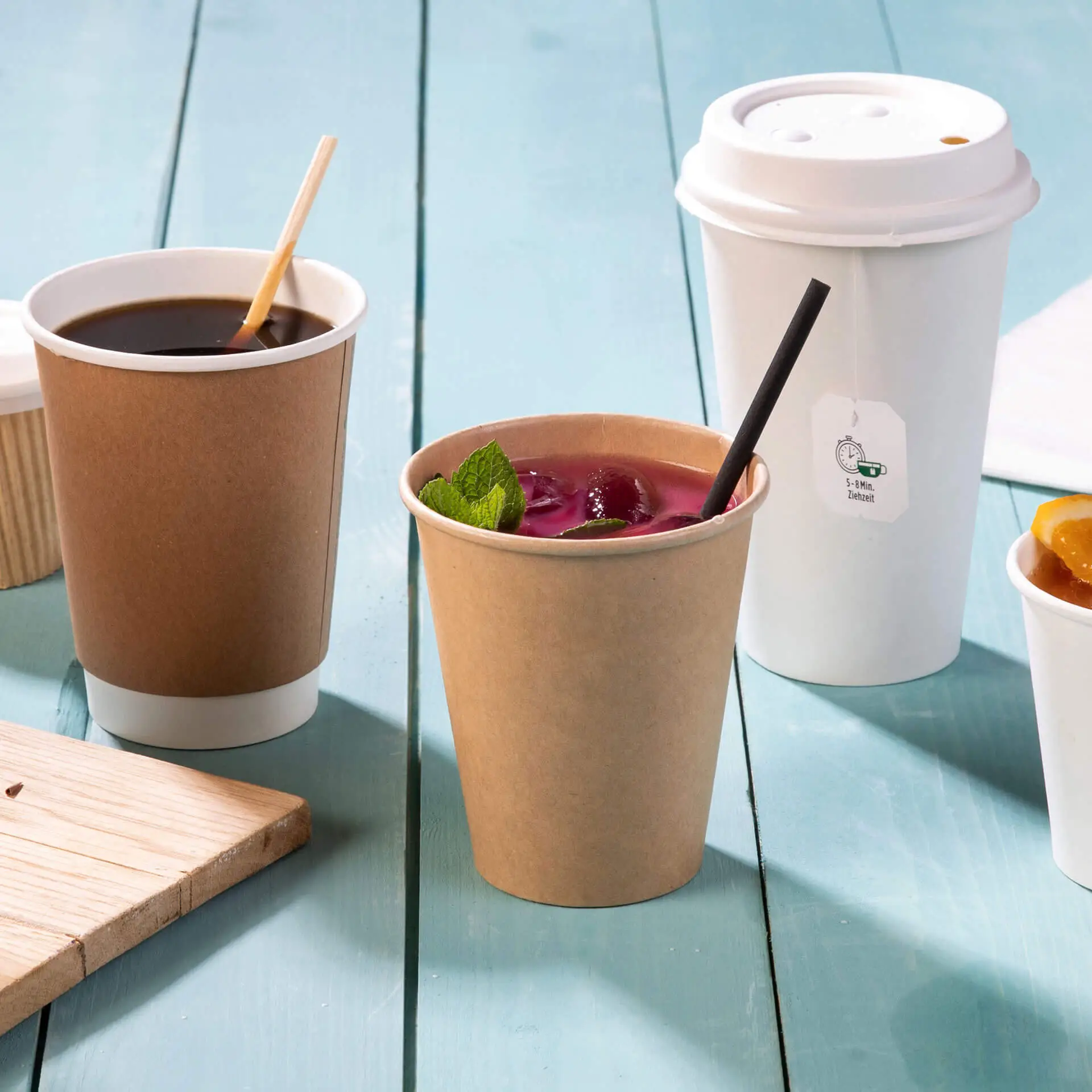 Un bicchiere di carta marrone con una cannuccia e una bevanda colorata, circondato da una tazza di caffè e altri oggetti su un tavolo azzurro.