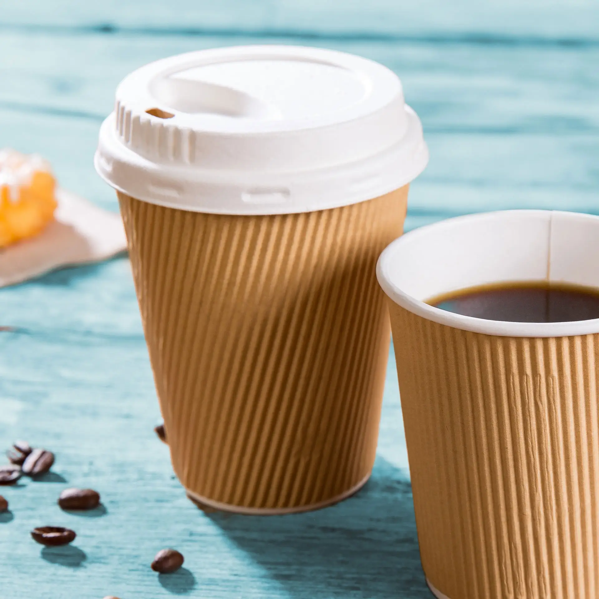 Due tazze da caffè da asporto marroni a ondulazione, una con coperchio, su una superficie turchese; chicchi di caffè sparsi vicino.