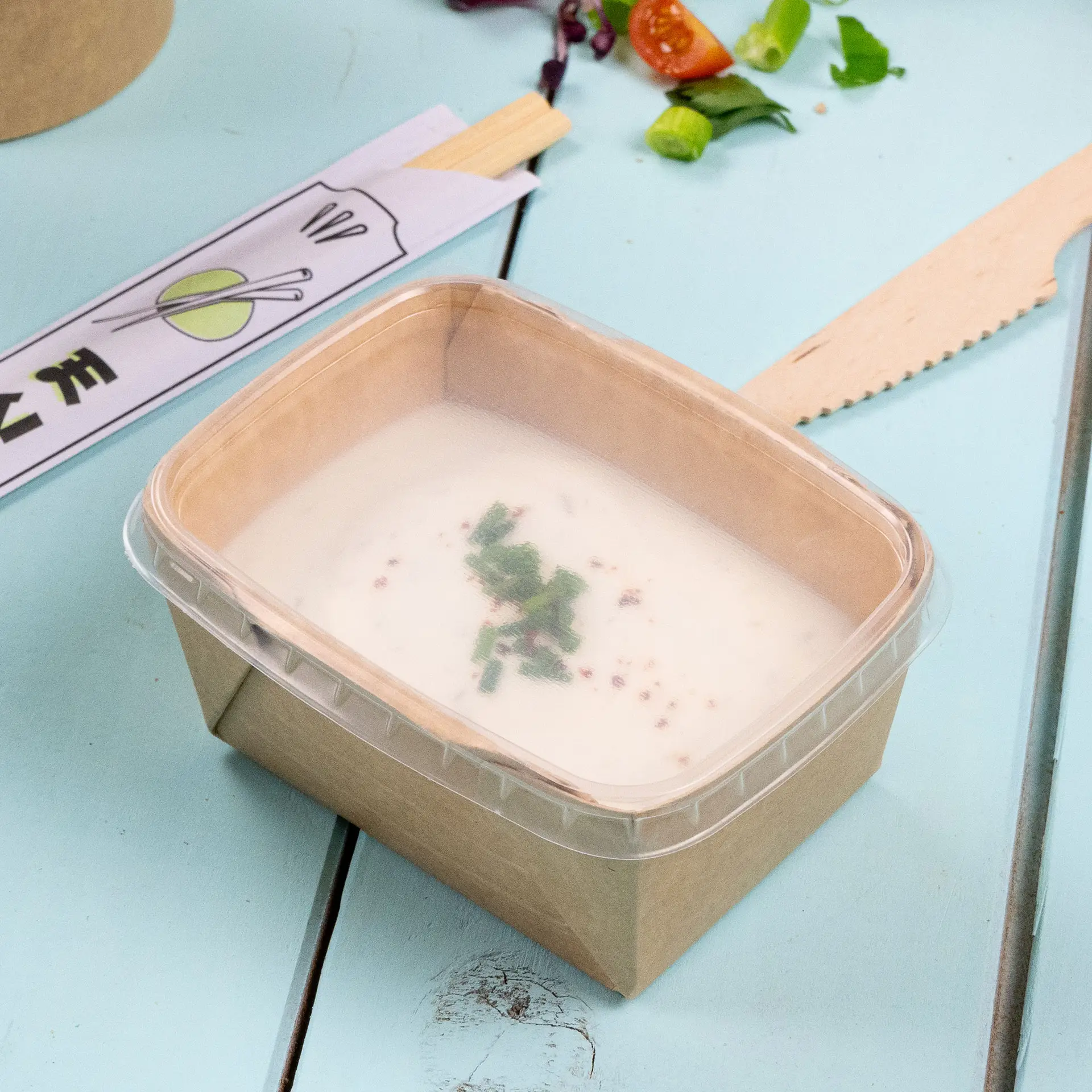 Coperchio PP trasparente su una ciotola rettangolare in cartone con zuppa cremosa e erbe, posata su una superficie blu chiaro.