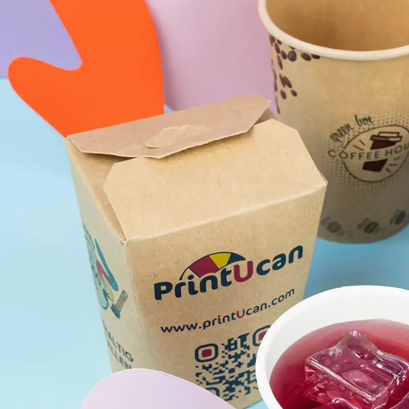 Contenitori di cartone marrone per ramen noodles con logo, posizionati su una superficie blu con vari condimenti e bacchette.