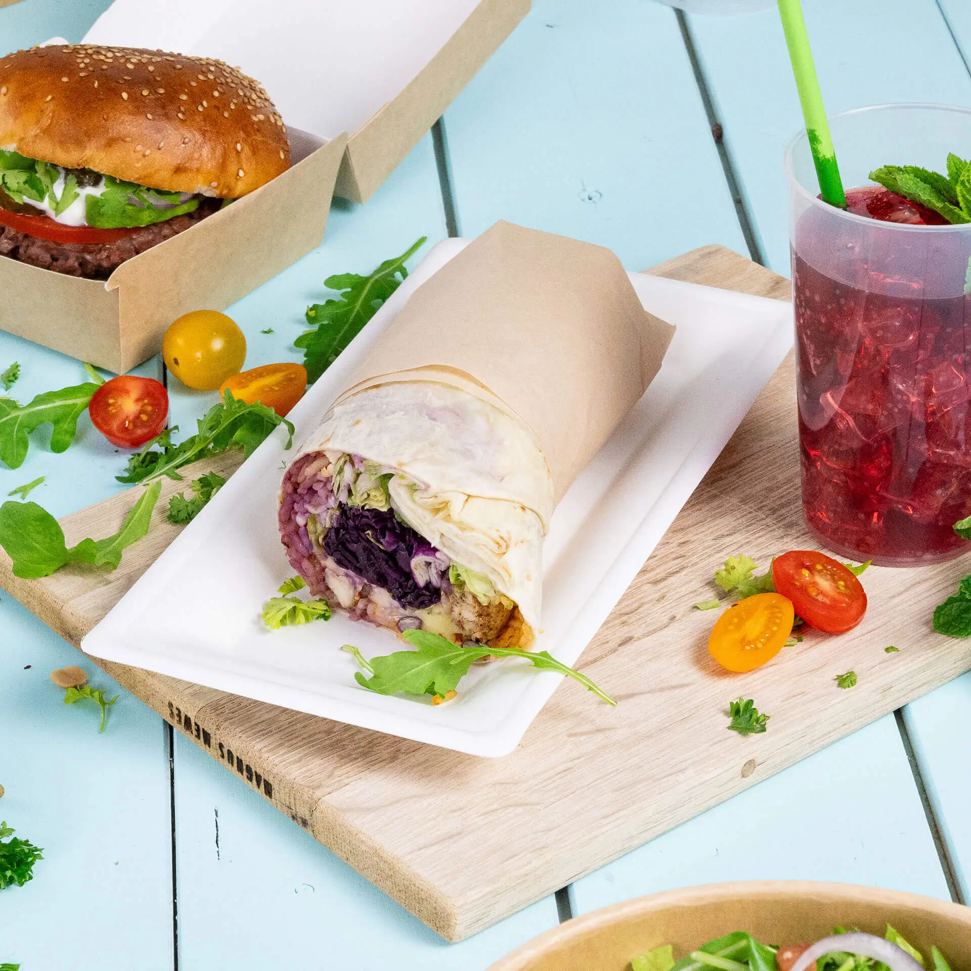 Piatto rettangolare in canna da zucchero con un involtino di verdure, un hamburger, pomodorini e una bevanda.