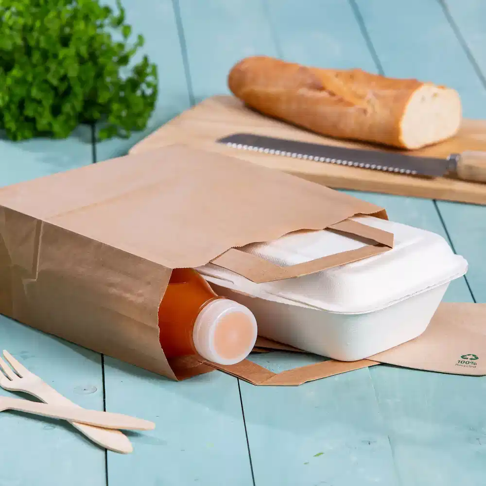 Sacchetto di carta marrone contenente una bottiglia di bevanda e un contenitore per cibo da asporto su una superficie in legno blu.