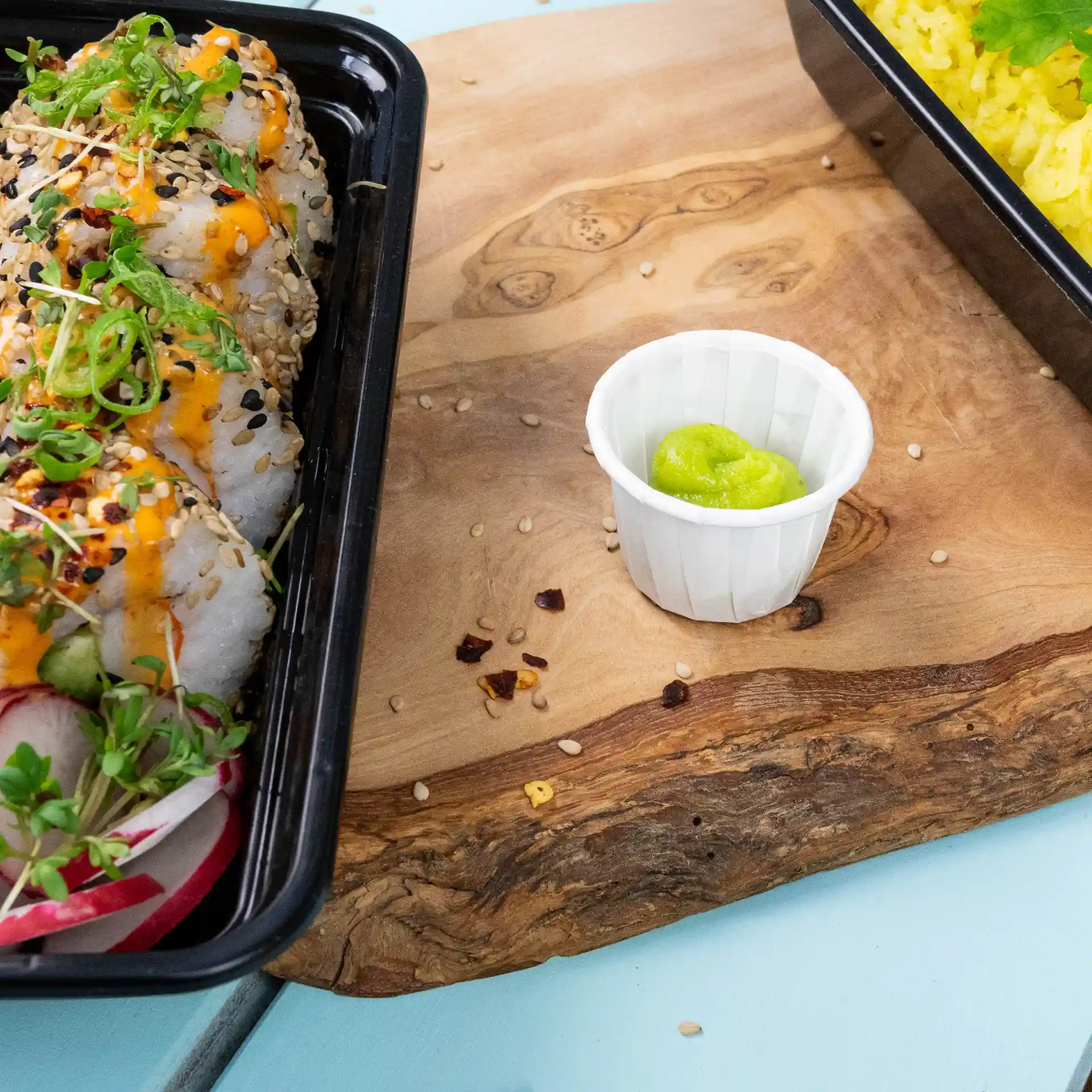 Rolls di sushi con semi di sesamo e guarnizione verde accanto a un piccolo bicchiere di carta bianco contenente wasabi su una superficie di legno.
