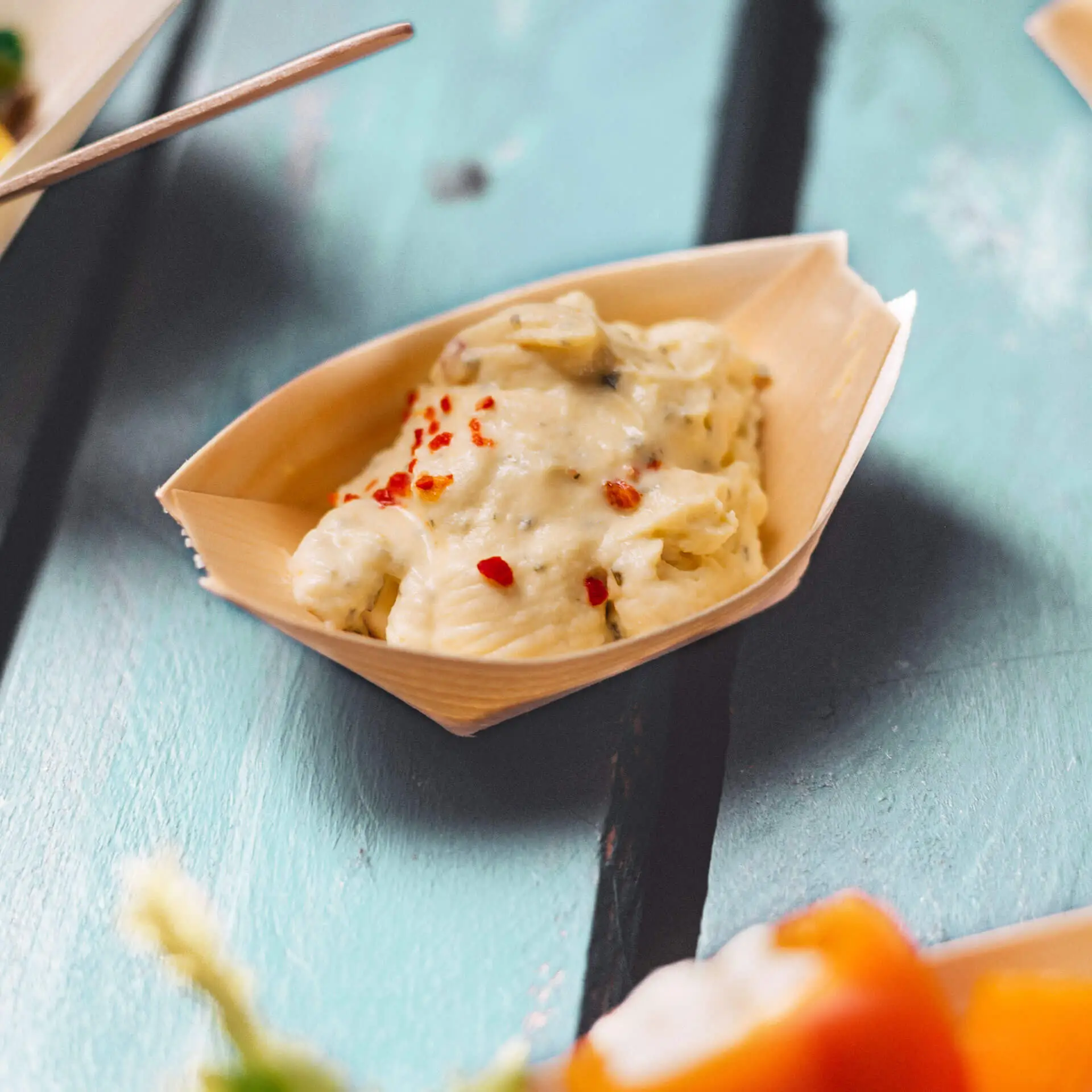 Ciotola in legno a forma di barca contenente cibo cremoso guarnito con fiocchi rossi, posata su una superficie in legno turchese.
