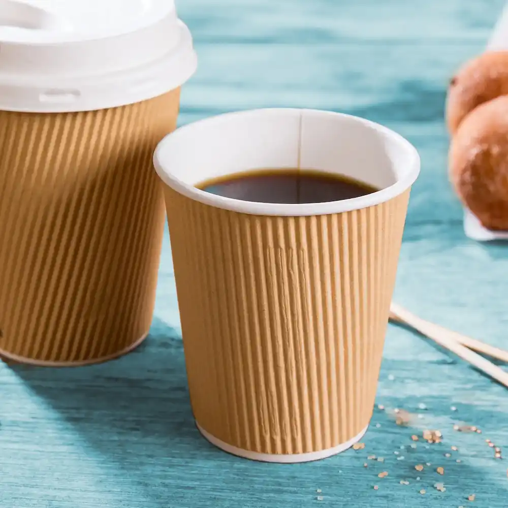 Bicchiere di carta marrone a coste con caffè nero, accanto a un bicchiere simile e ciambelle su una superficie blu.