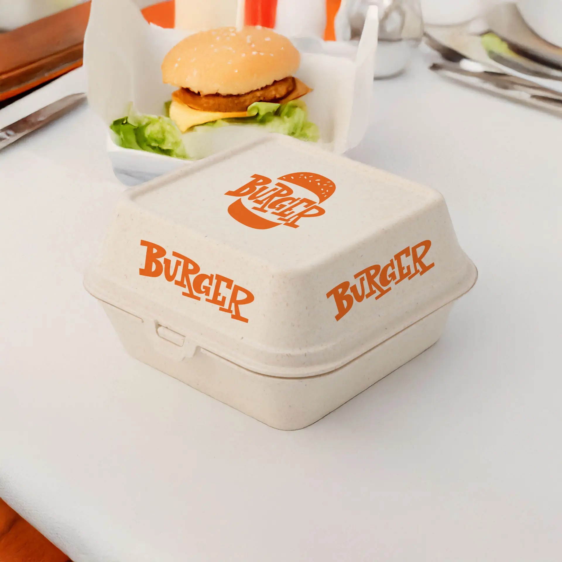 Scatola per hamburger rettangolare bianca crema con testo stampato arancione, posizionata su un tavolo accanto a un hamburger e lattuga.