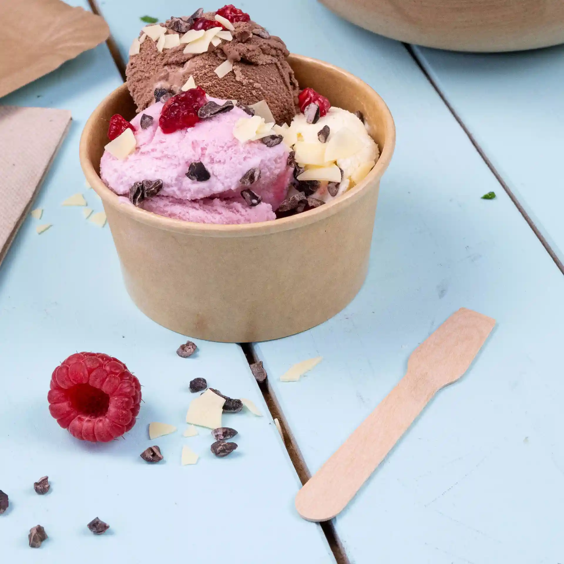 Becher con tre palline di gelato rosa, marrone e bianco, con guarnizioni, accanto a un cucchiaio di legno e un lampone.