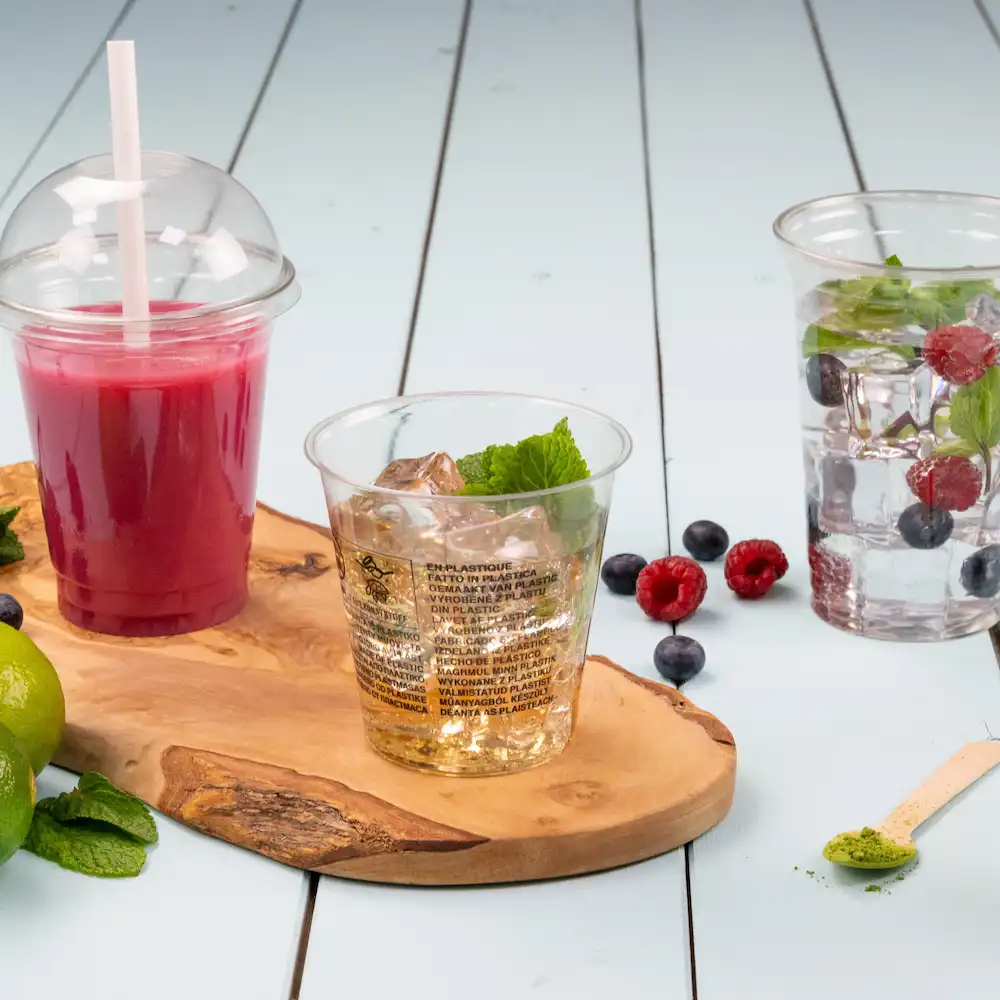 Bicchieri di plastica trasparenti con bevande e menta su un tagliere di legno, con frutti di bosco e lime sparsi attorno.