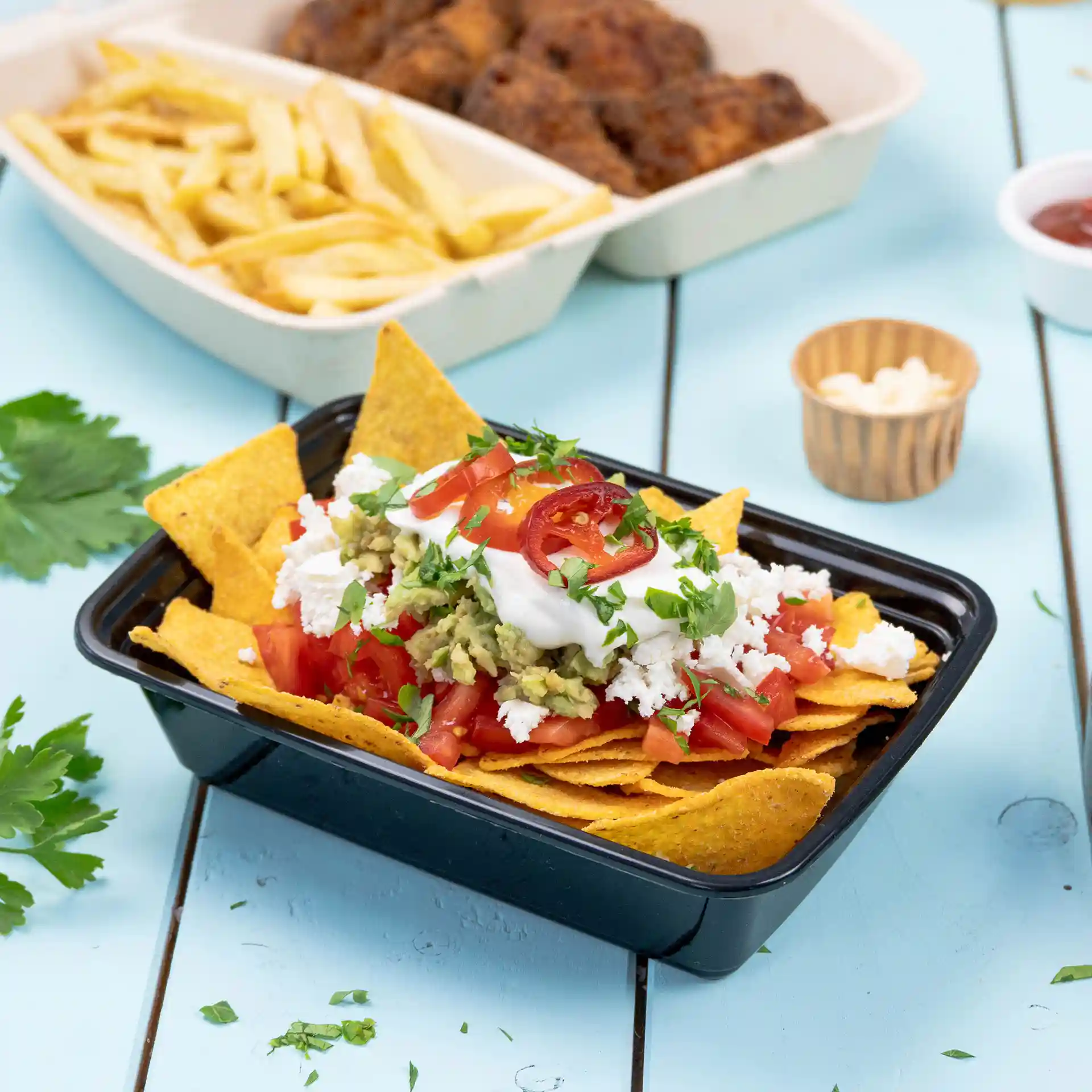 Contenitore rettangolare riutilizzabile nero riempito di nachos, guarnito con pomodori, guacamole, panna acida e coriandolo.