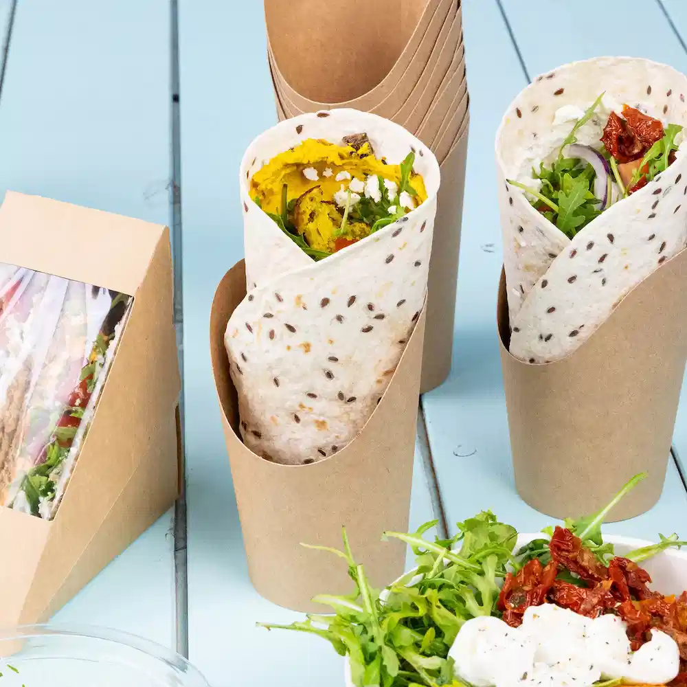 Due wrap in contenitori di carta con ingredienti visibili, insieme a un'insalata e pomodori secchi su un tavolo.