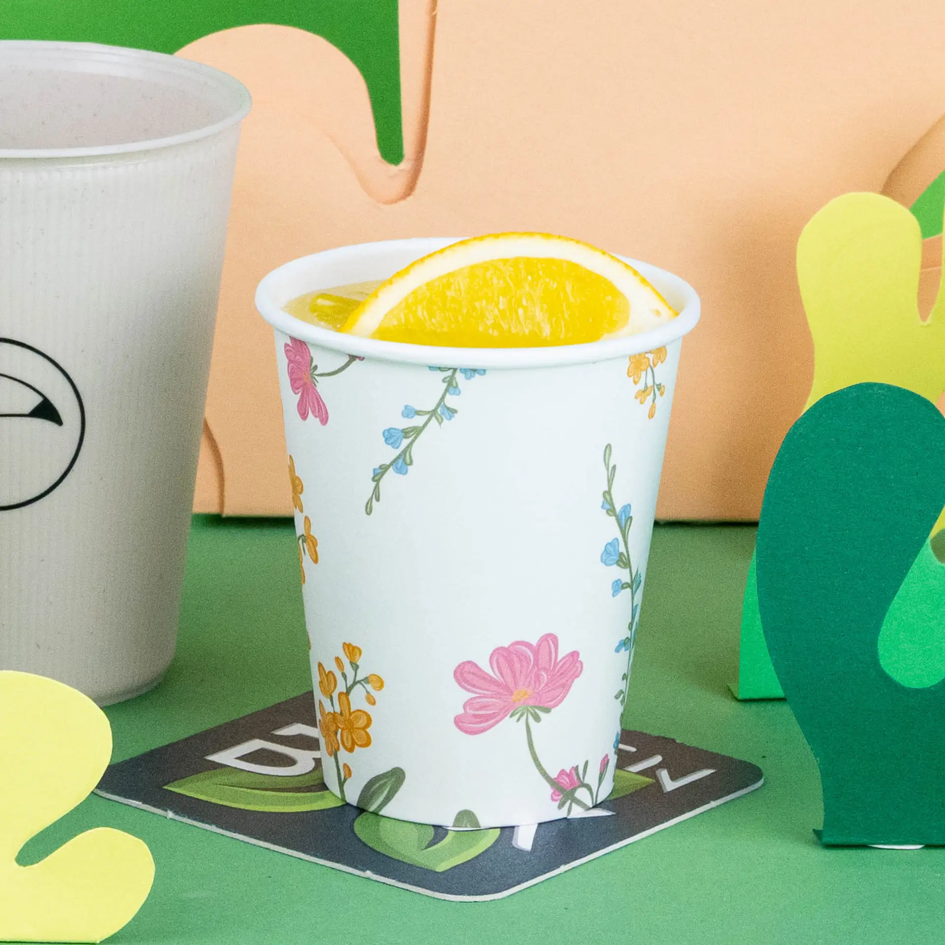 Bicchiere bianco con design floreale, riempito di una bevanda e guarnito con una fetta di limone, su sfondo verde.