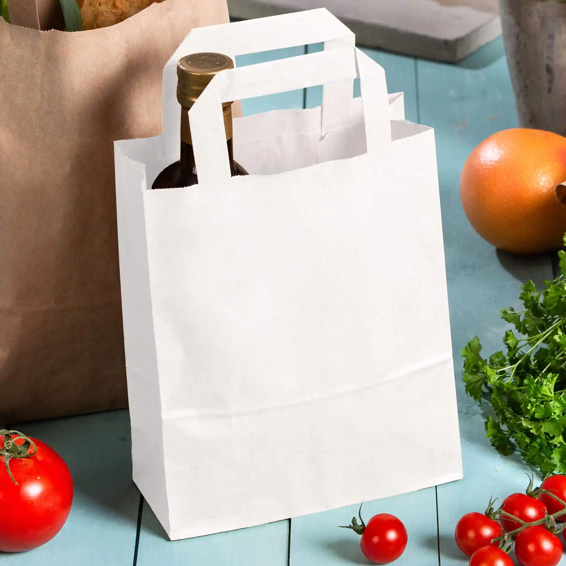 Borsa in carta kraft bianca con manici, che tiene una bottiglia, circondata da pomodori e un'arancia su una superficie blu.