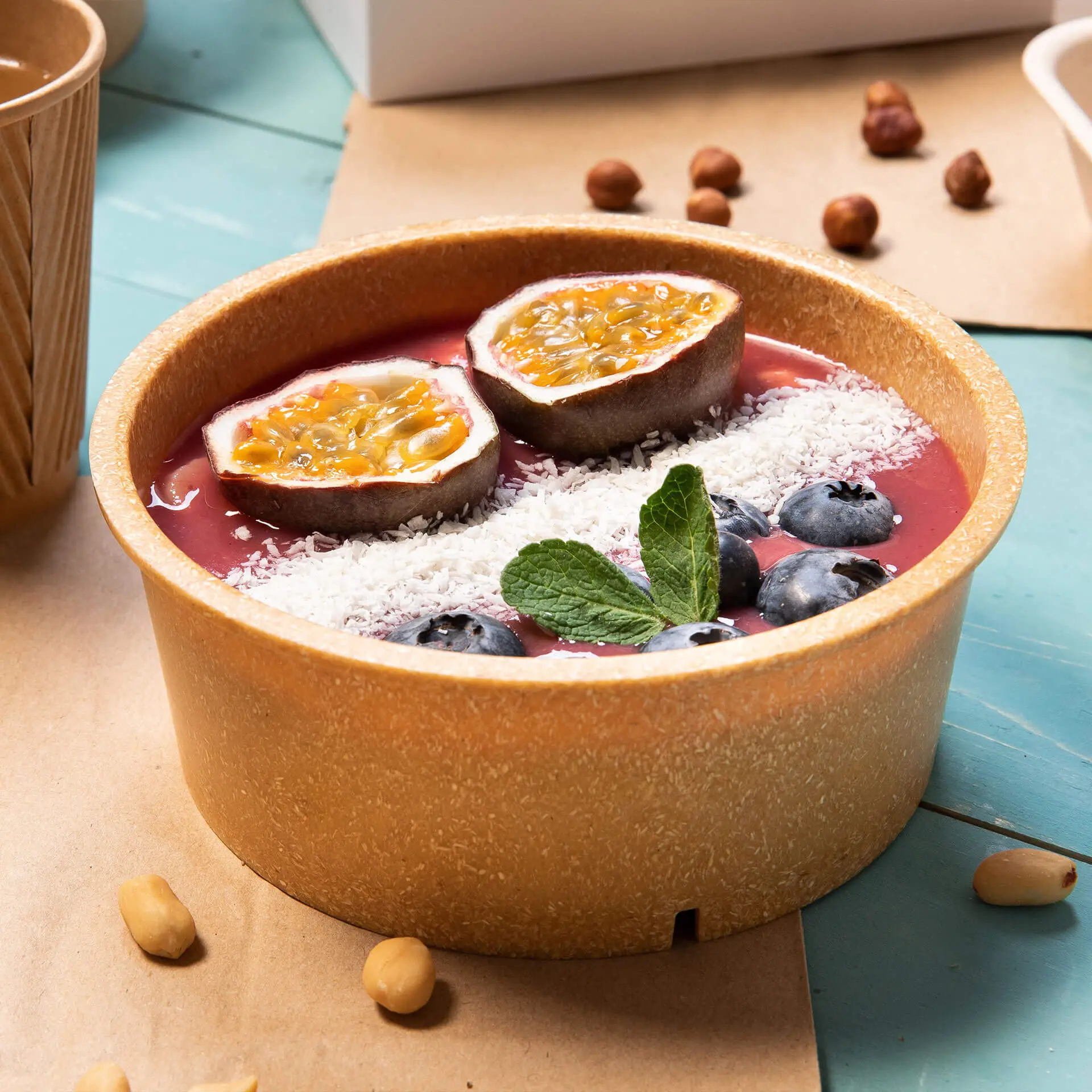 Una ciotola marrone riutilizzabile piena di smoothie rosa, guarnita con metà frutto della passione, cocco grattugiato, mirtilli e menta.