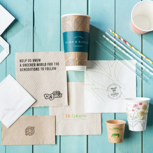 Tovaglioli di carta marroni e bianchi, bicchieri usa e getta e cannucce su una superficie di legno blu.