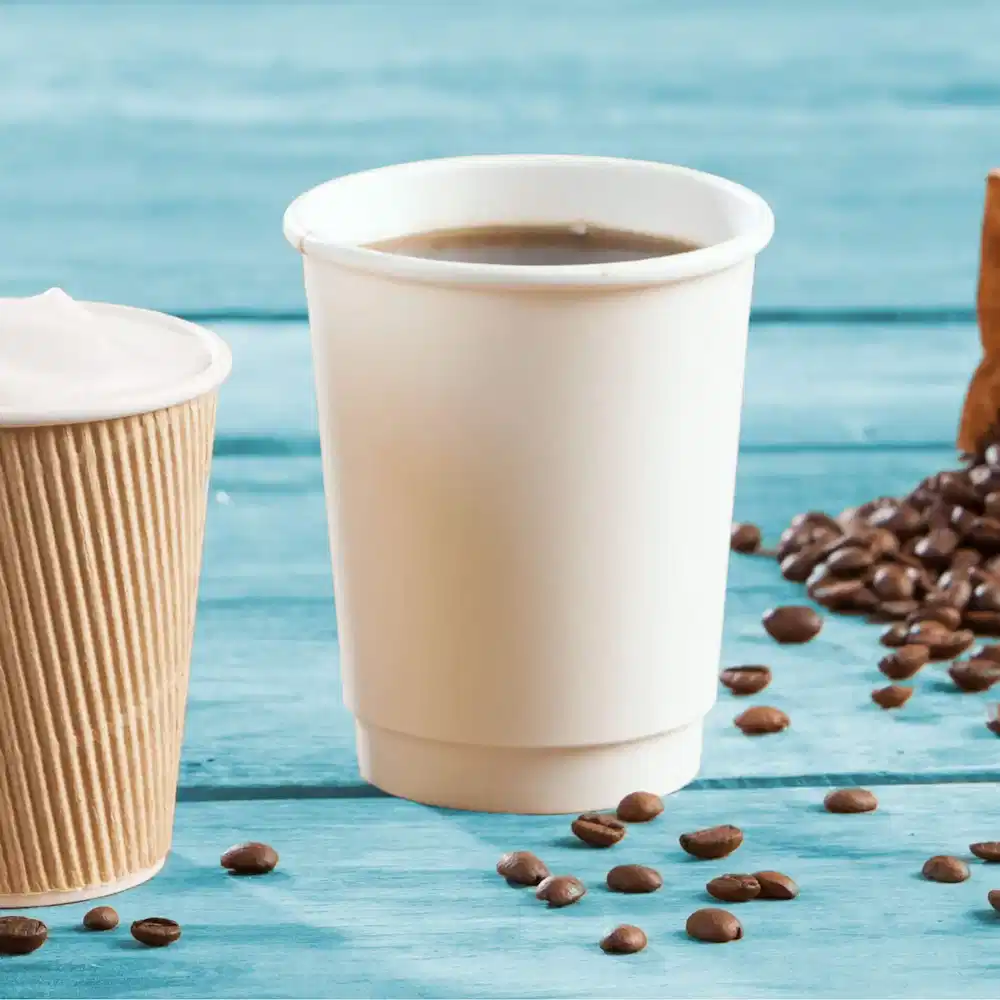 Due bicchieri di carta a doppia parete impilati, uno bianco semplice e uno marrone rigato, con chicchi di caffè sparsi.
