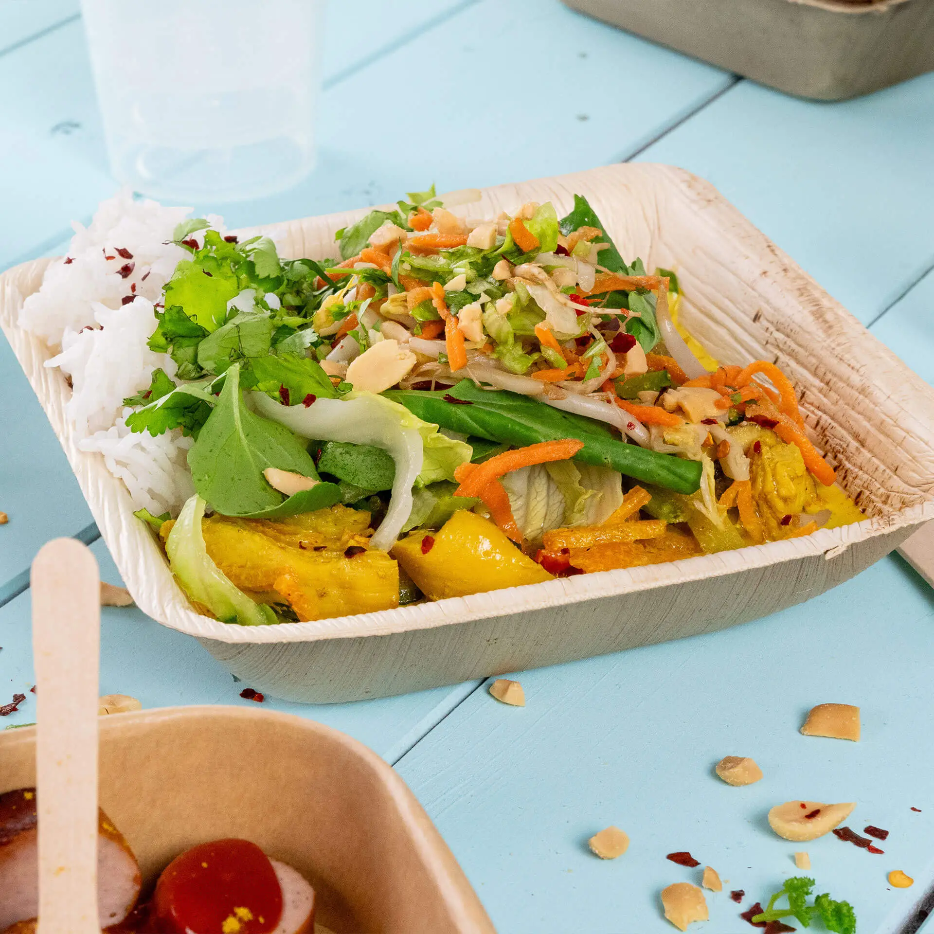 Una ciotola di foglie di palma piena di riso, insalata e verdure, posata su una superficie azzurra chiara.