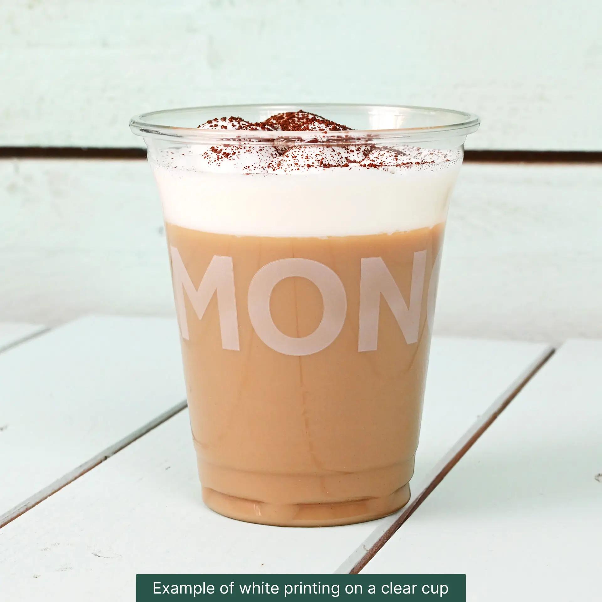 Bicchiere rPET trasparente ripieno di caffè ghiacciato, guarnito con panna montata e cacao, su un piano in legno.