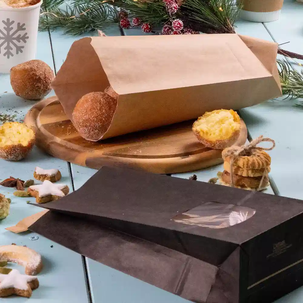 Sacchetto di carta marrone con prodotti da forno, borsa nera con finestra trasparente, tagliere in legno e decorazioni festive.
