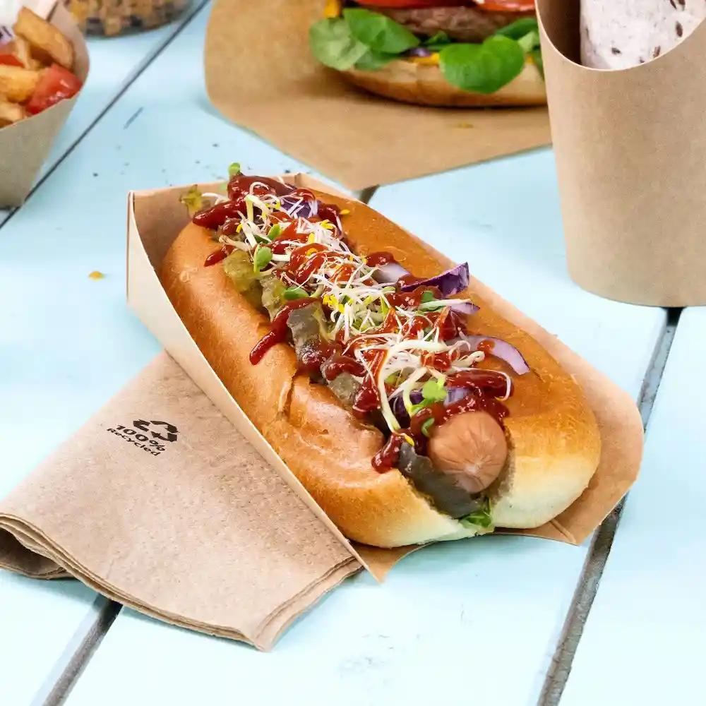 Hot dog in un portacibo di carta marrone, guarnito con ketchup, senape, cetriolini e germogli, poggiato su un tavolo di legno.