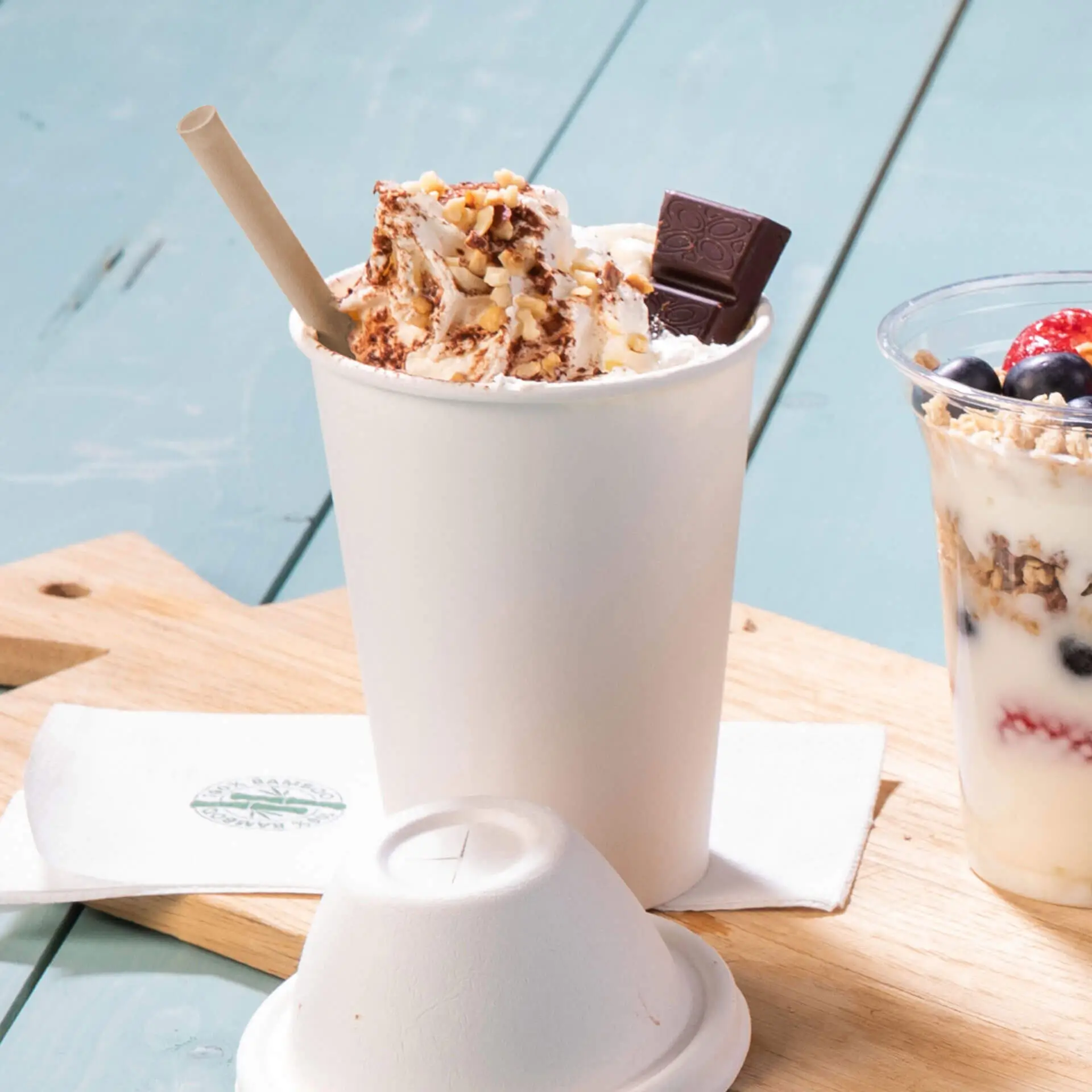 Un bicchiere di carta bianco riempito di panna montata, guarnito con noci e un pezzo di cioccolato, accanto a un bicchiere trasparente di parfait di yogurt.