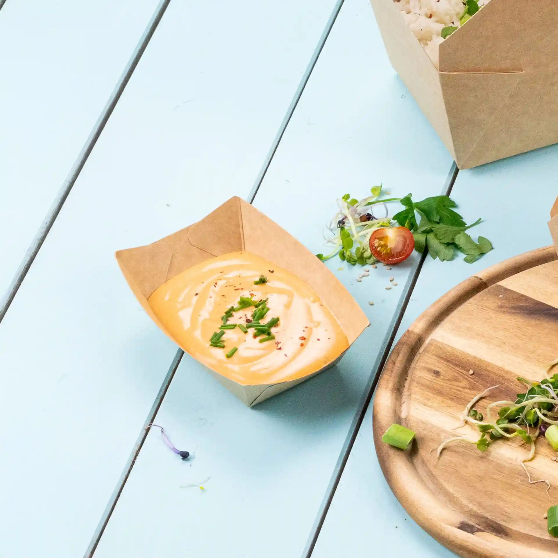 Vassoio per snack in cartone marrone con una salsa cremosa guarnita con erba cipollina tritata, posato su una superficie azzurro chiaro.