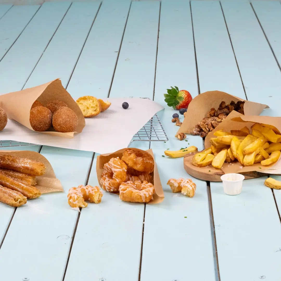 Varietà di cibi fritti e dolci serviti in involucri di carta su una superficie in legno blu chiaro.