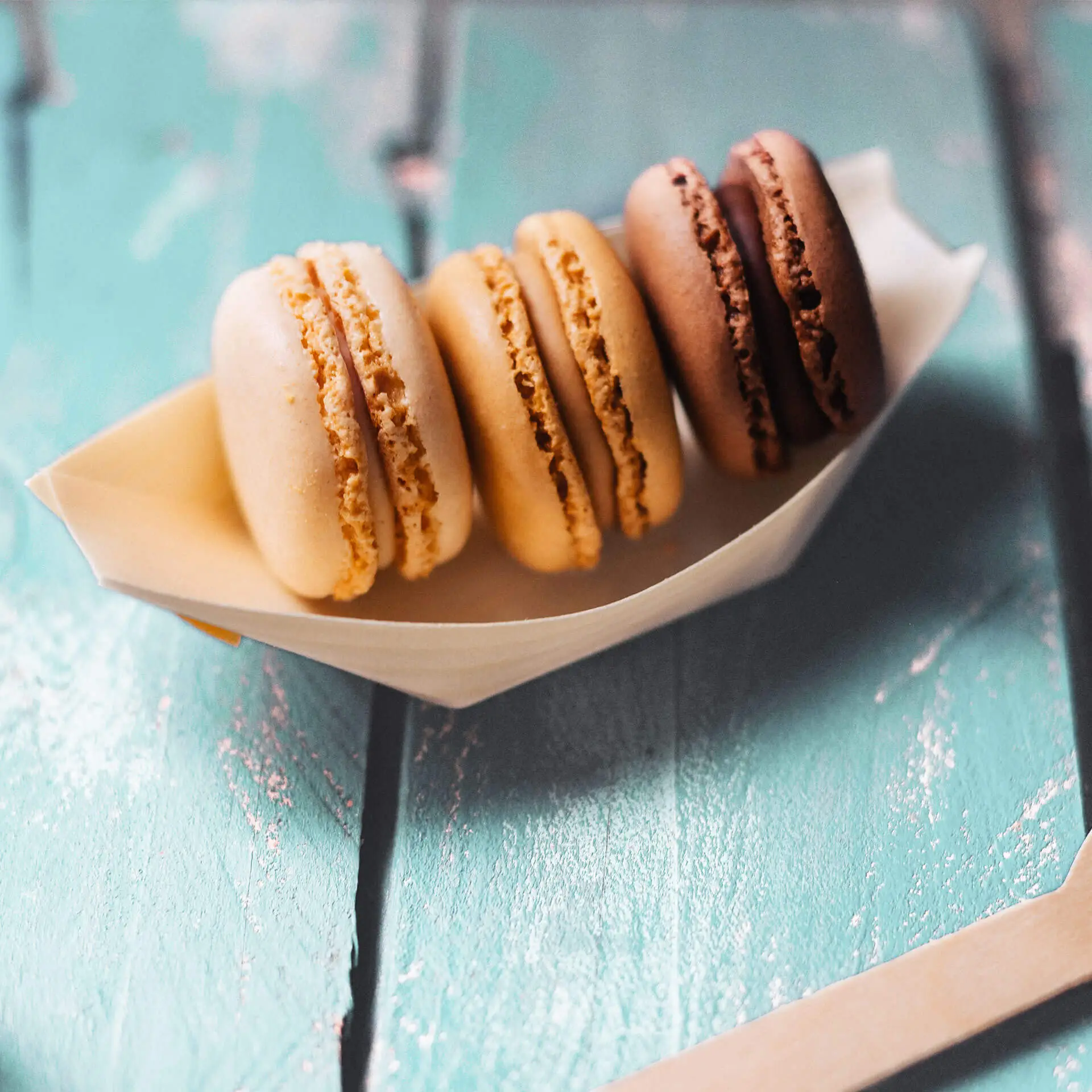 Quattro macarons in una barchetta di legno decorativa, con colori beige chiaro, dorato, marrone medio e marrone scuro, disposti ordinatamente.