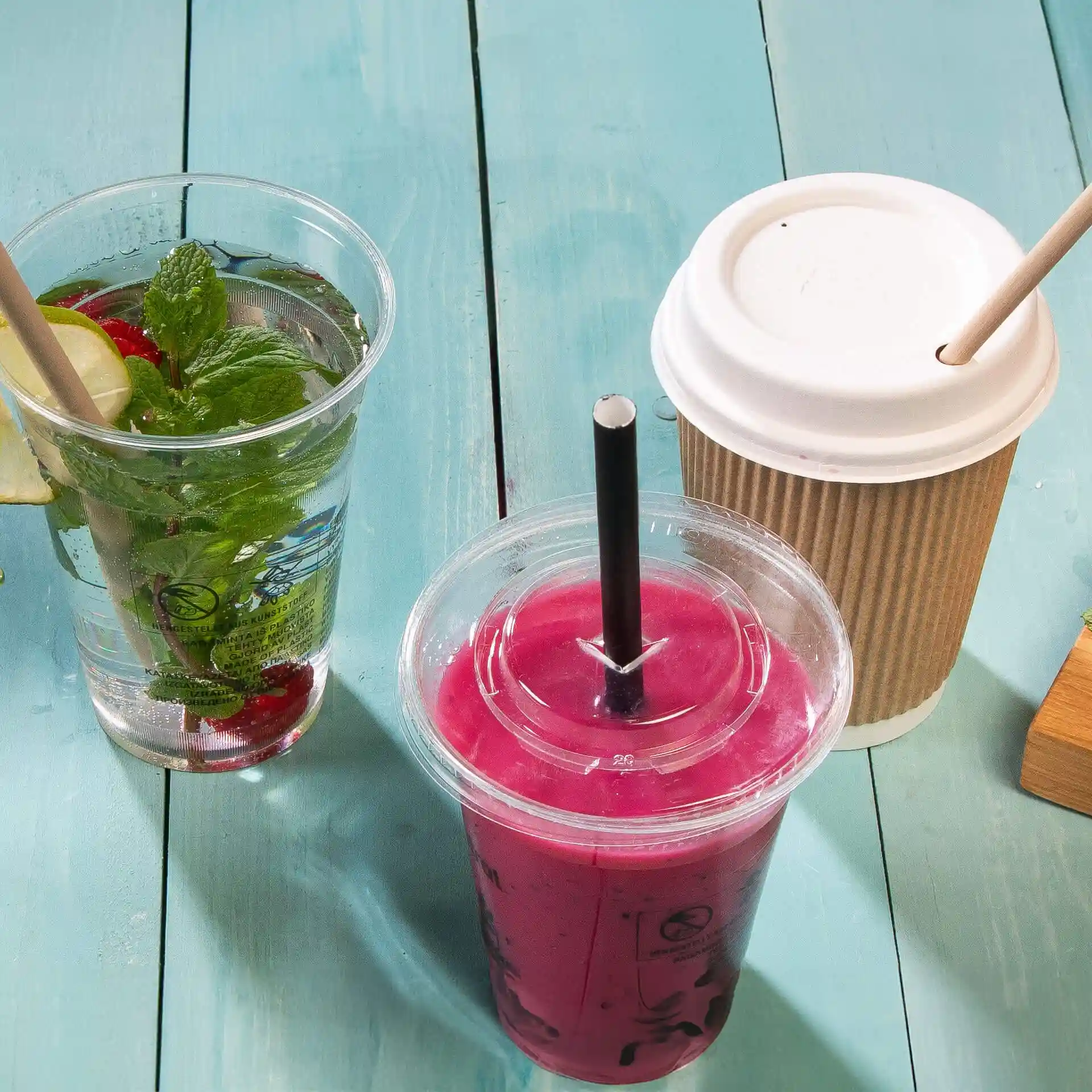 Tre bevande su una superficie di legno blu: un bicchiere trasparente con menta e limone, uno smoothie rosa con una cannuccia nera e un bicchiere di carta marrone con un coperchio bianco.