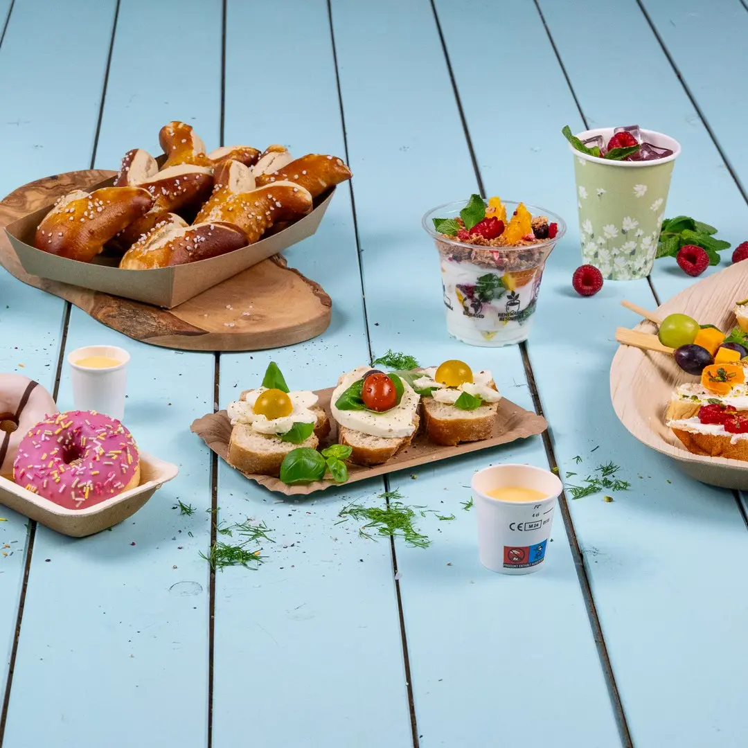 Snack assortiti tra cui pretzel, panini con condimenti, parfait di yogurt, ciambelle e spiedini di frutta su una superficie di legno blu.