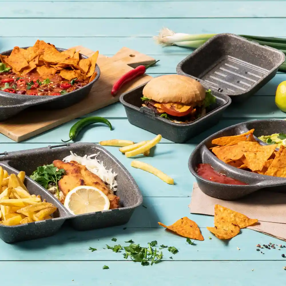 Varietà di cibi in contenitori riutilizzabili, tra cui nachos, un hamburger, patatine e pesce con limone, su una superficie blu.
