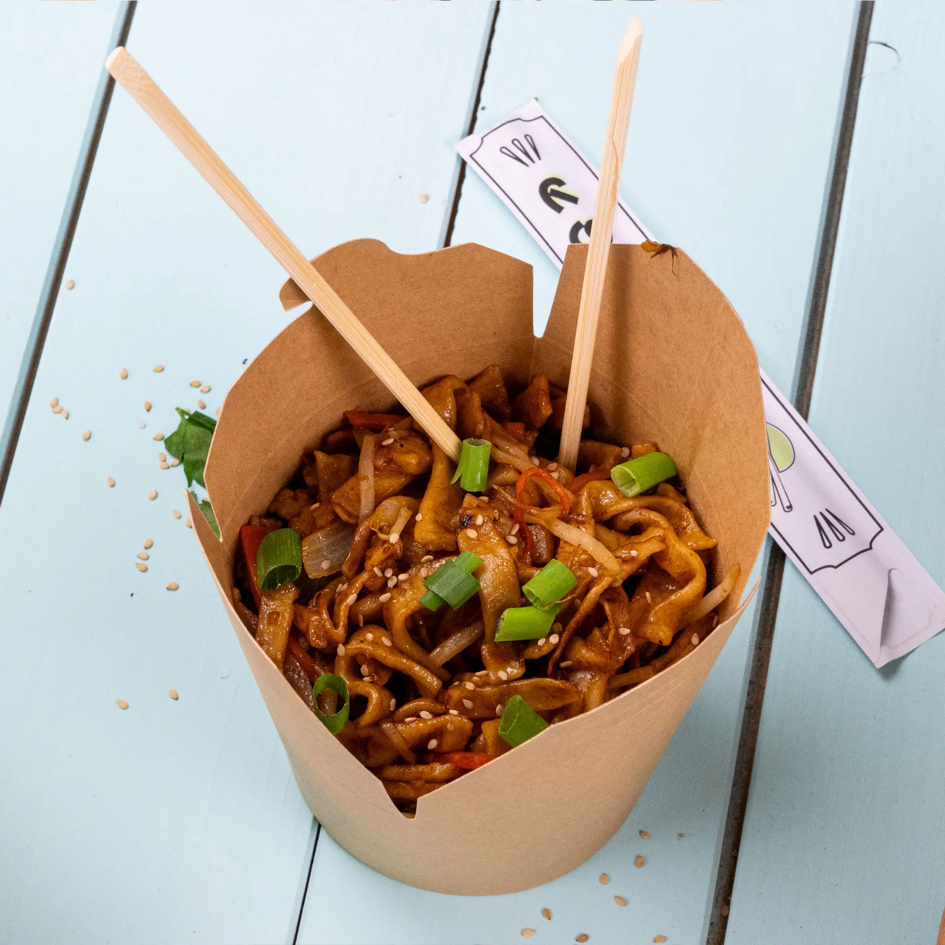 Bacchette di bambù in un contenitore di carta riempito con noodle saltati, guarniti con cipollotti e semi di sesamo.