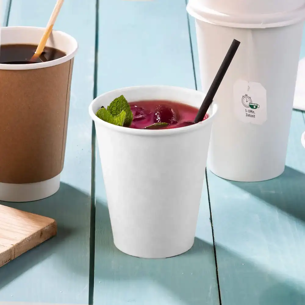 Bicchiere di carta bianco contenente una bevanda con foglie di menta, una cannuccia nera e due altri bicchieri sullo sfondo.