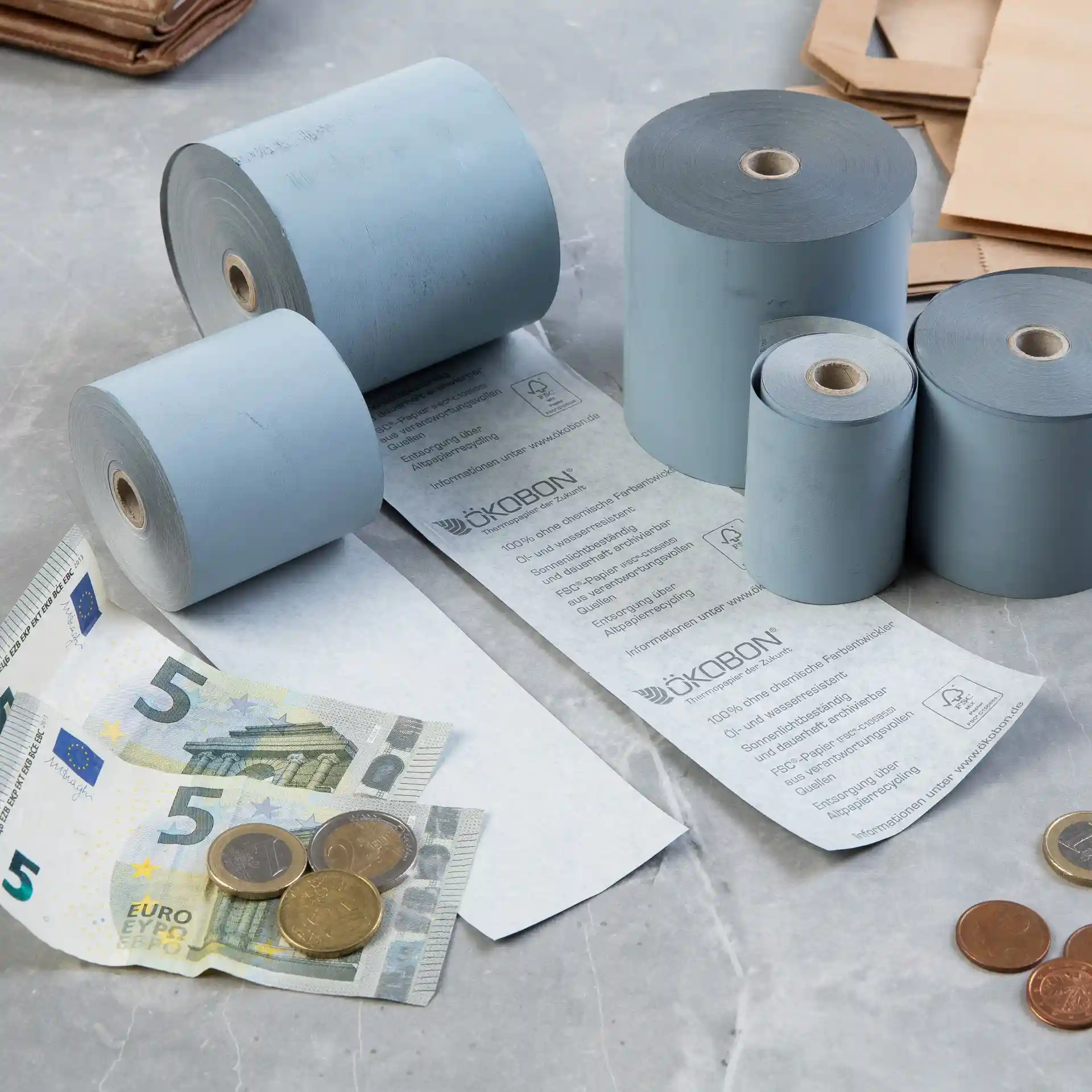 Immagine di diversi rotoli di carta termica grigia, una banconota da 5 euro e monete su una superficie.