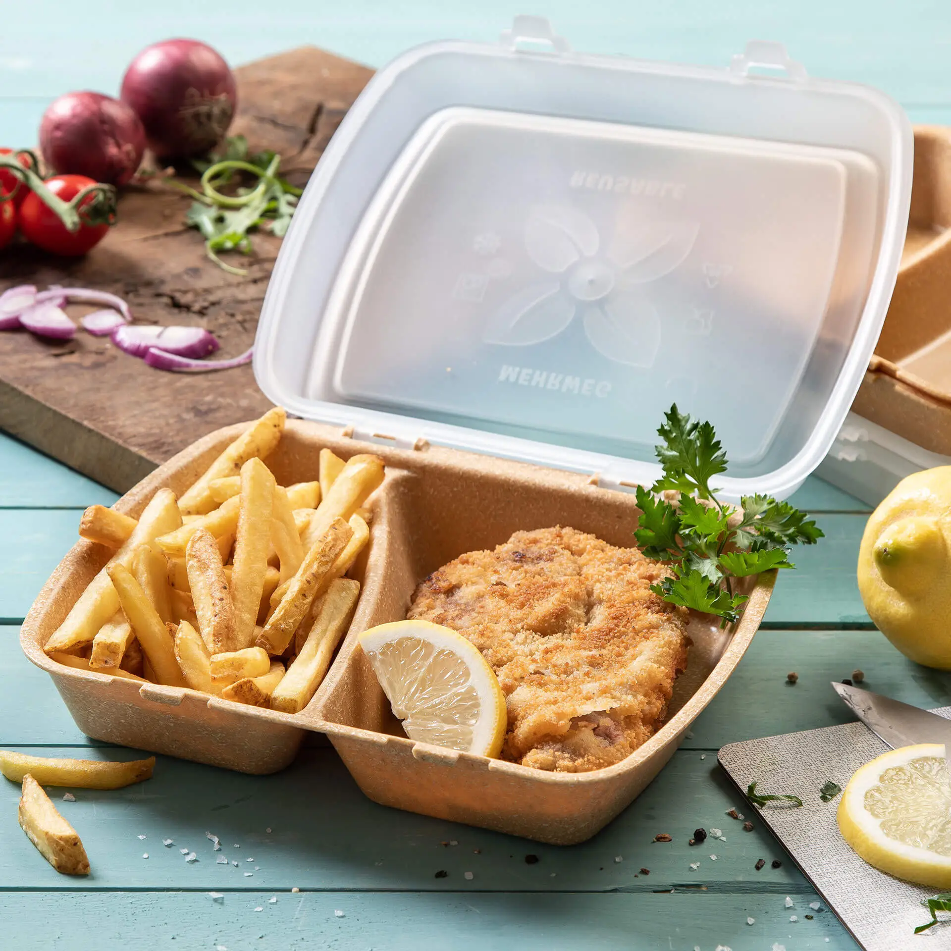 Contenitore con pollo fritto, patatine e fetta di limone, in una scatola di plastica trasparente su una superficie blu.