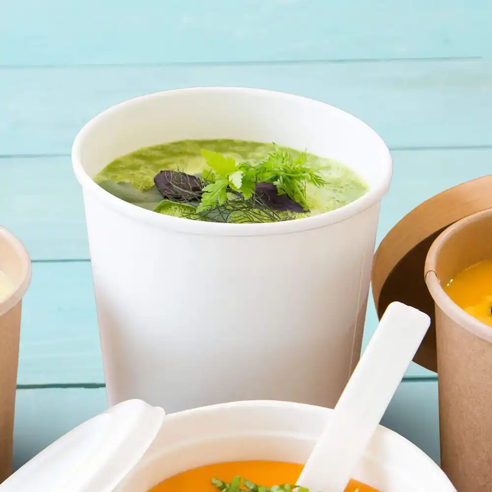 Tazza bianca per zuppa riempita di zuppa verde e guarnita con erbe, circondata da altri contenitori.