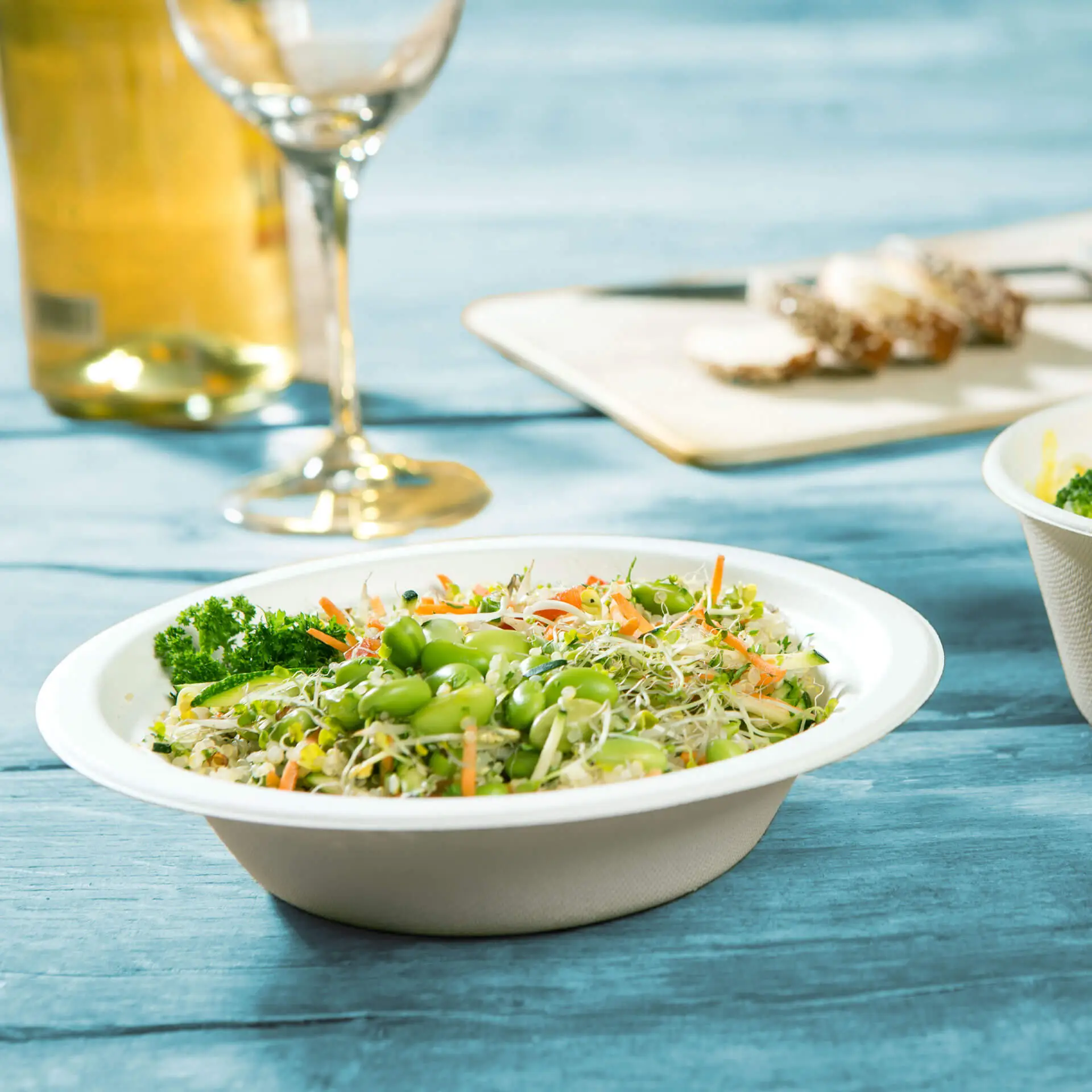 Una ciotola piena di un'insalata di verdure fresca, con edamame e germogli, su un tavolo di legno blu, con un bicchiere di vino sullo sfondo.