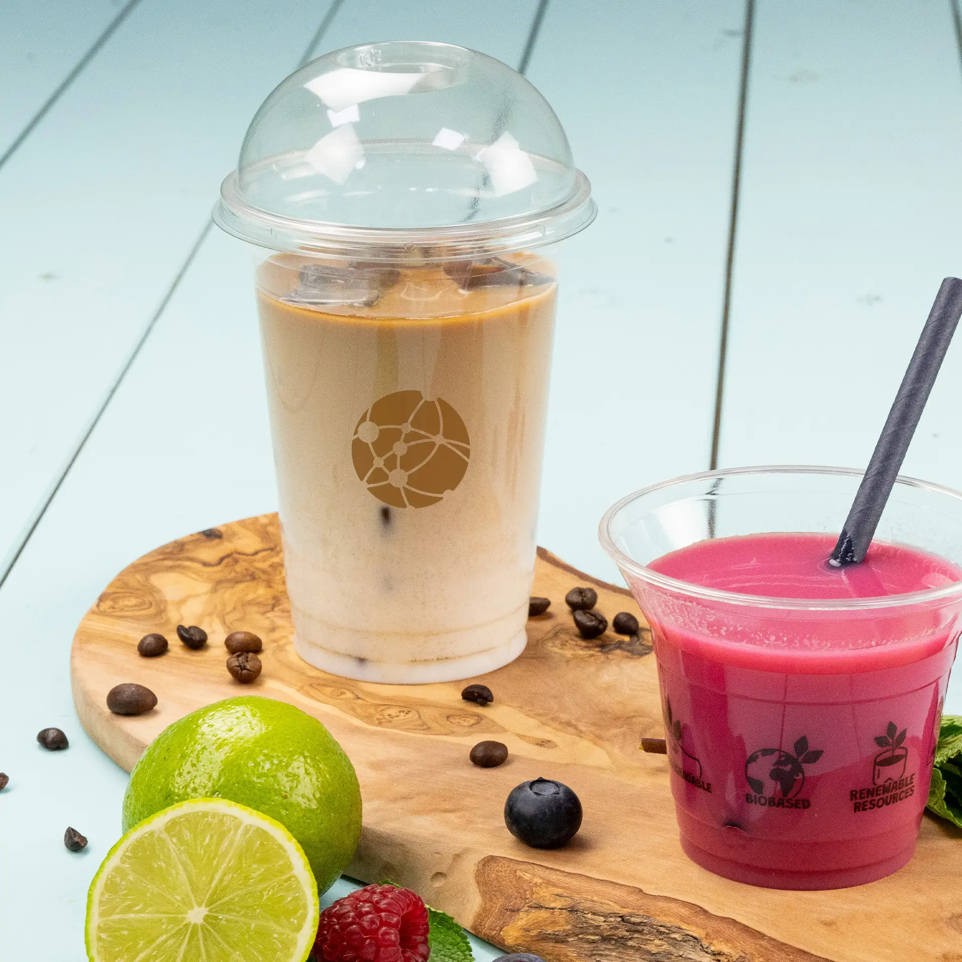 Un bicchiere di PLA trasparente riempito di caffè freddo, etichettato con un logo; accanto, un bicchiere più piccolo con una bevanda rosa e una cannuccia, su una tavola di legno.