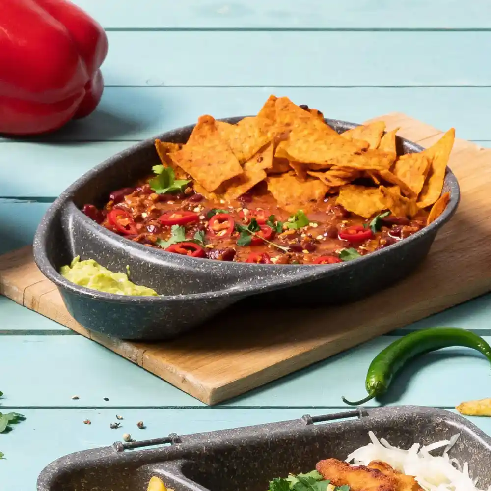 Ciotola di chili guarnita con nachos, peperoncini rossi affettati e coriandolo su un tagliere di legno con condimenti.