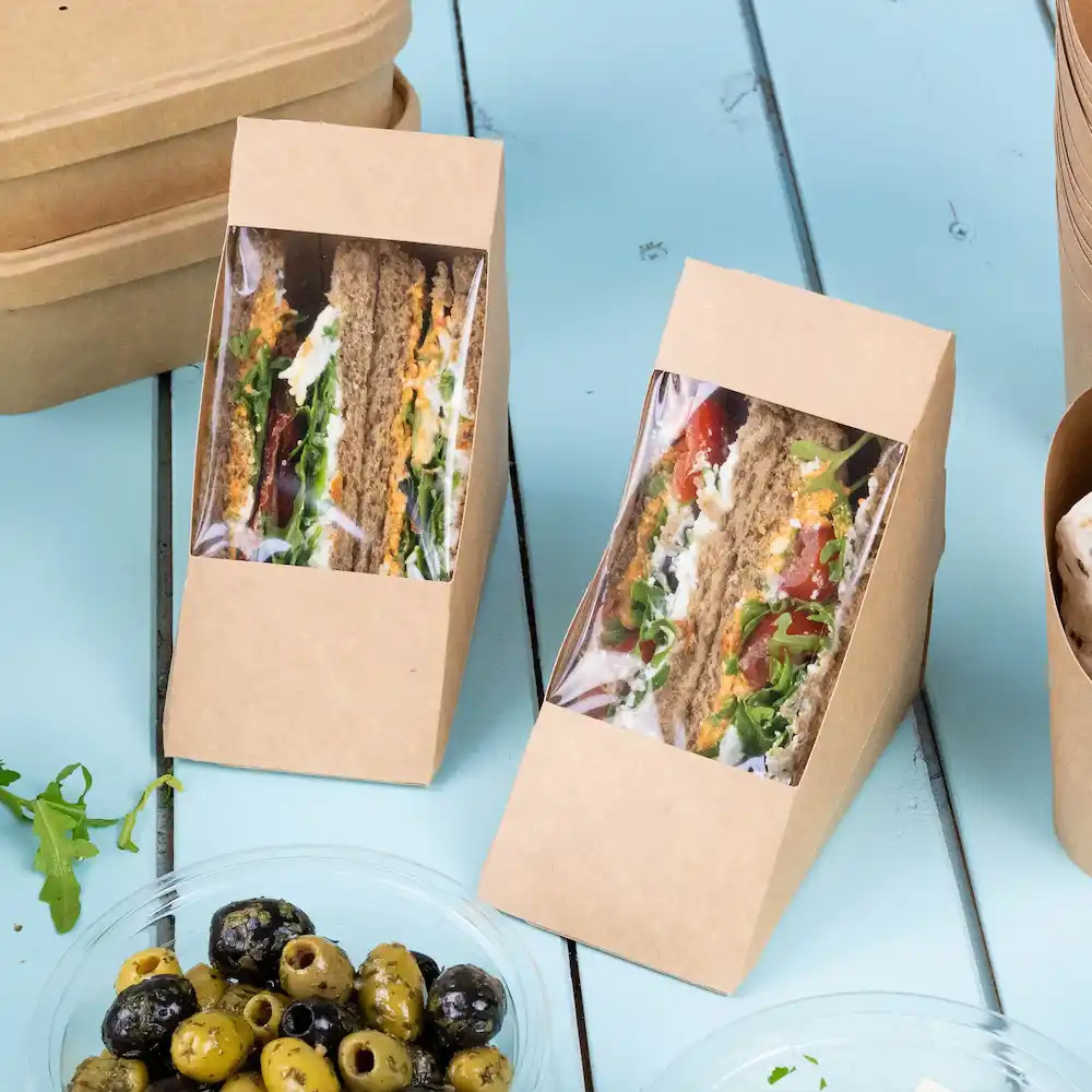 Due scatole per panini in carta kraft marrone con finestre che mostrano panini a strati e una ciotola di olive miste.