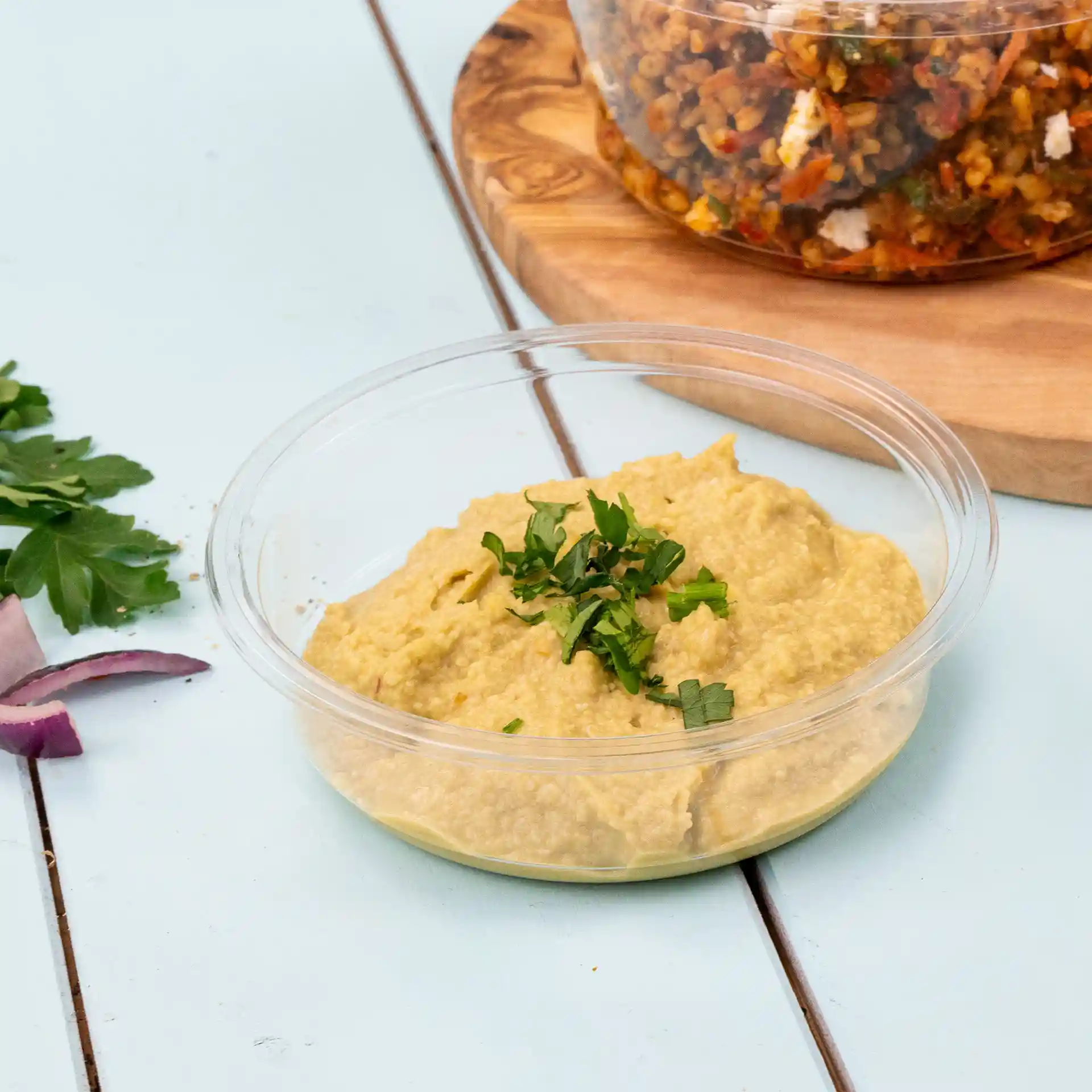 Contenitore deli trasparente rotondo con hummus guarnito con prezzemolo tritato, posato su una superficie azzurro chiaro.