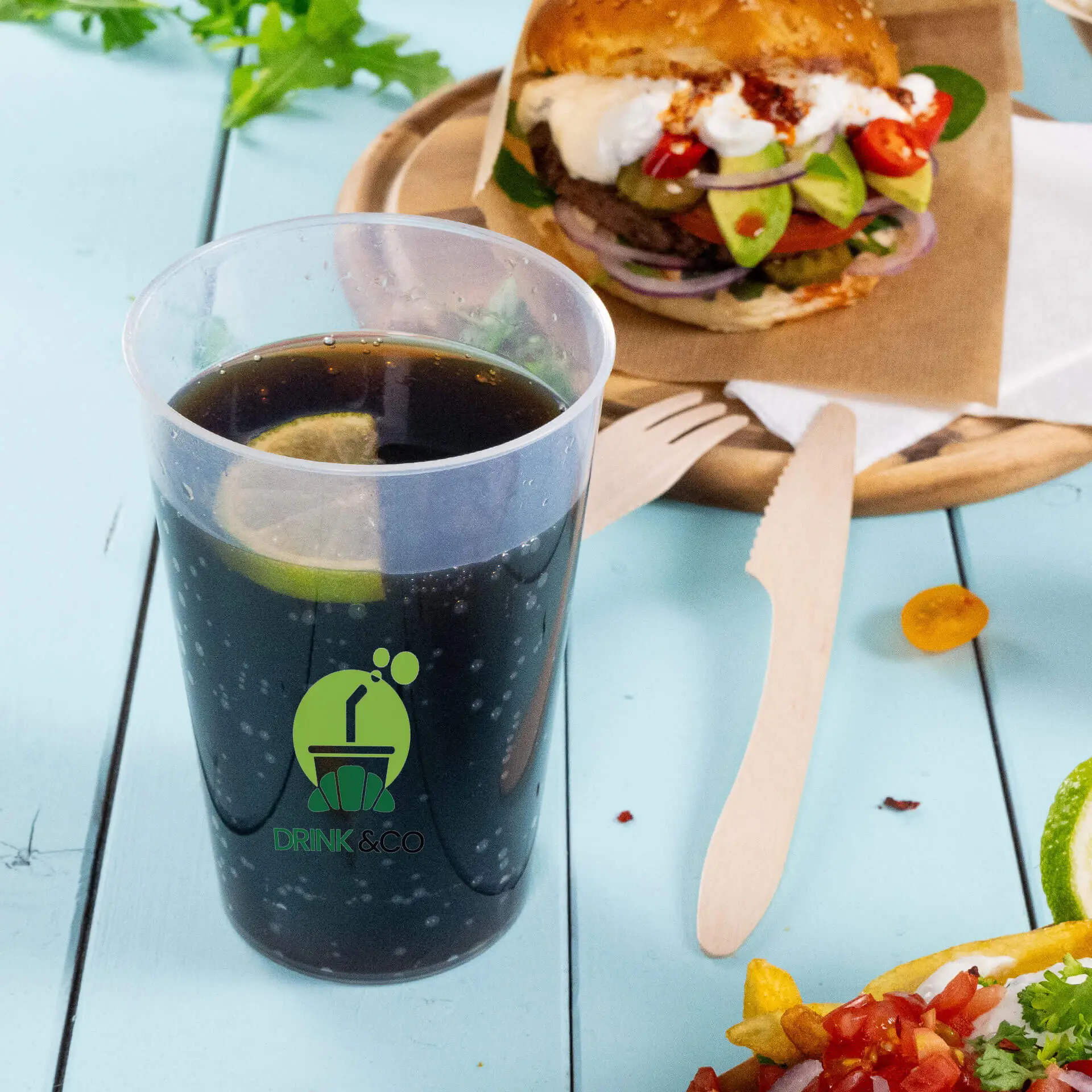 Un bicchiere riutilizzabile da 500 ml riempito con bevanda scura e una fetta di lime, con logo; hamburger e contorni sullo sfondo.