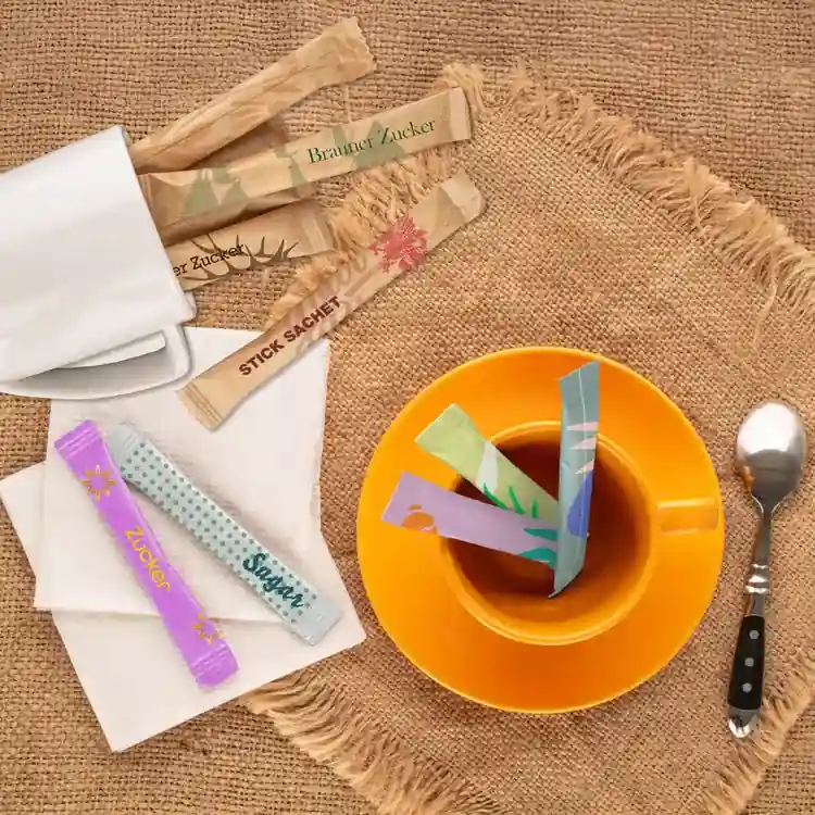 Sachet di zucchero assortiti posti in una tazza di tè su una superficie di juta, con un cucchiaio e un tovagliolo vicini.