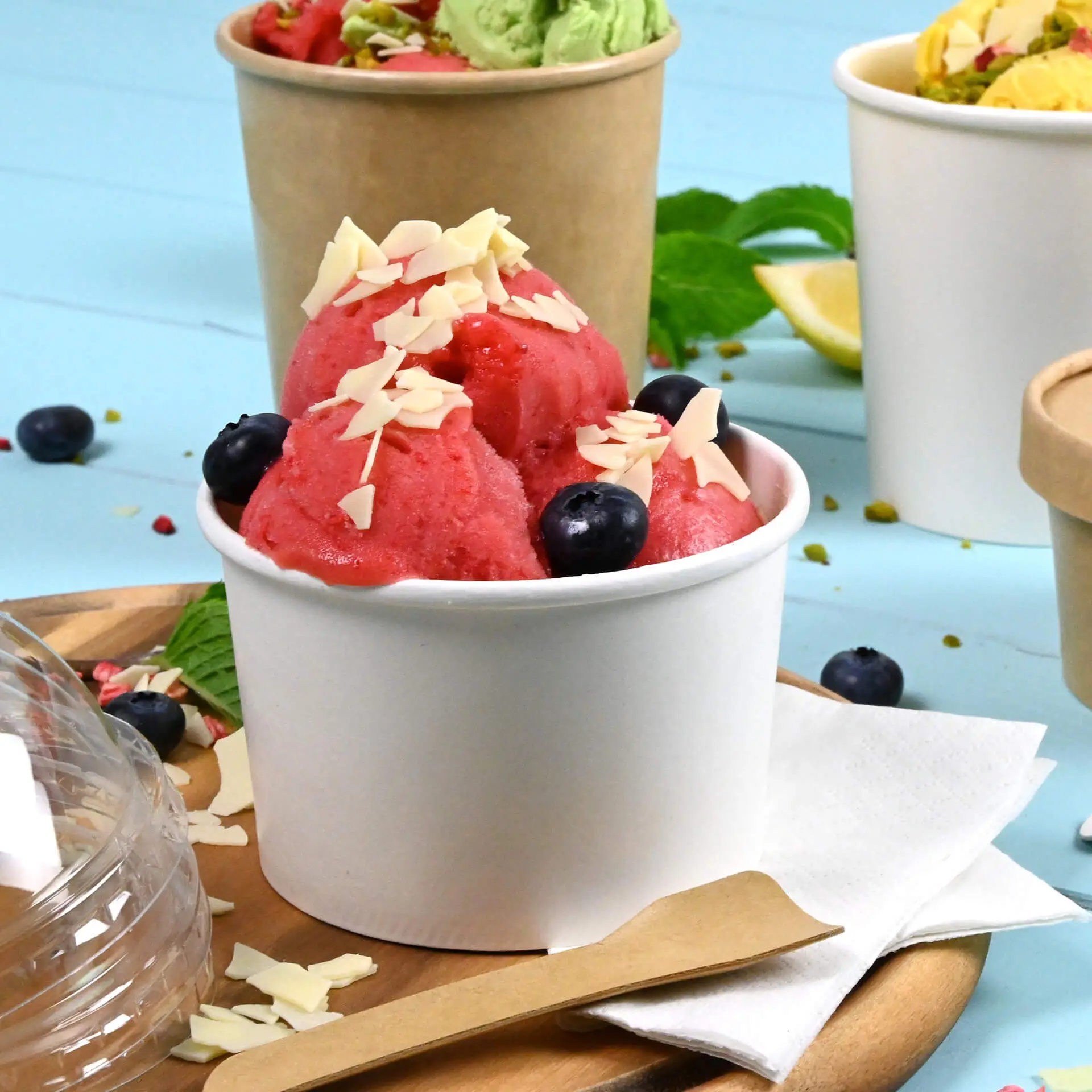 Un bicchiere bianco per gelato in PE pieno di sorbetto rosa, guarnito con scaglie di mandorla e mirtilli, su un vassoio di legno.