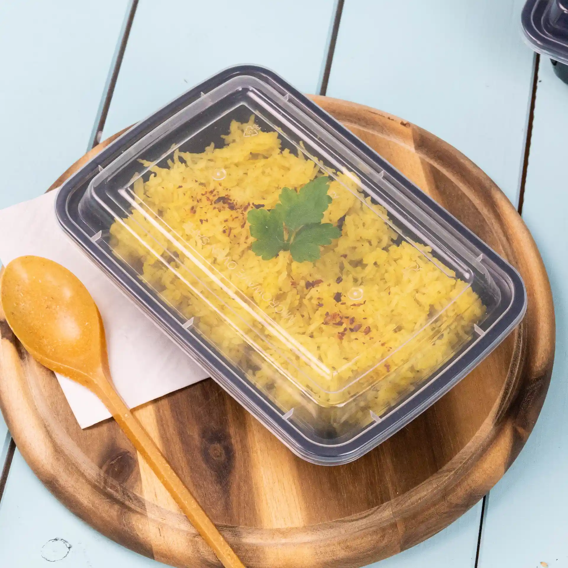 Contenitore rettangolare trasparente con coperchio riutilizzabile, contenente riso giallo e guarnito con una foglia di coriandolo, su un piatto di legno.