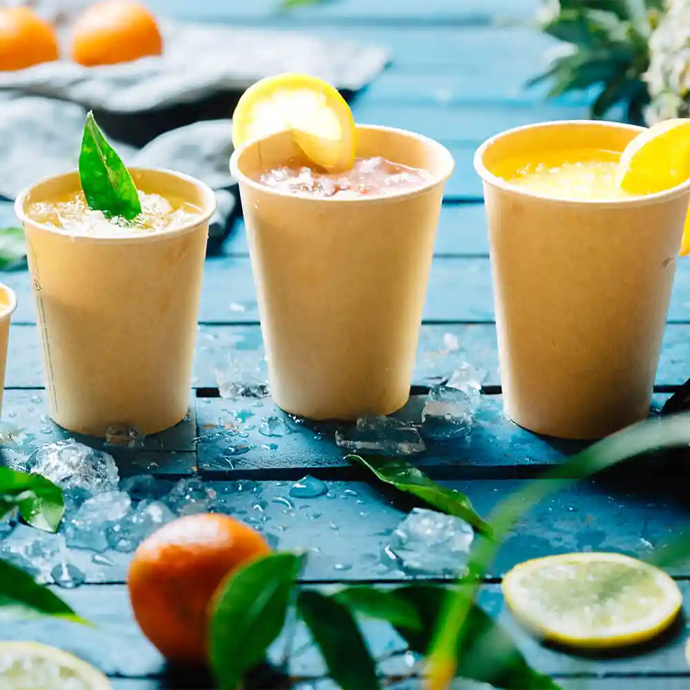 Tre bicchieri di carta marroni da 12 oz pieni di bevande, guarniti con limone e foglie, su una superficie di legno blu con ghiaccio.