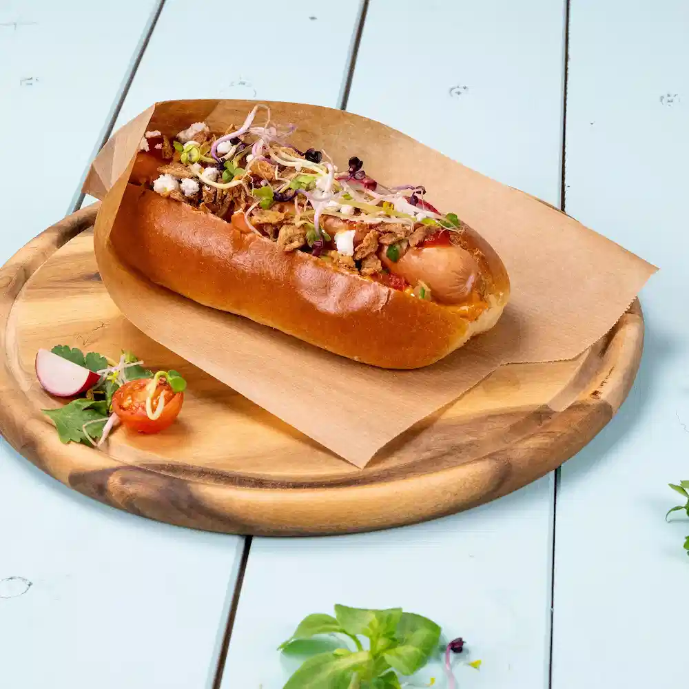 Hot dog in un panino guarnito con cipolle, erbe e condimenti croccanti su un vassoio di legno con contorni di verdure.